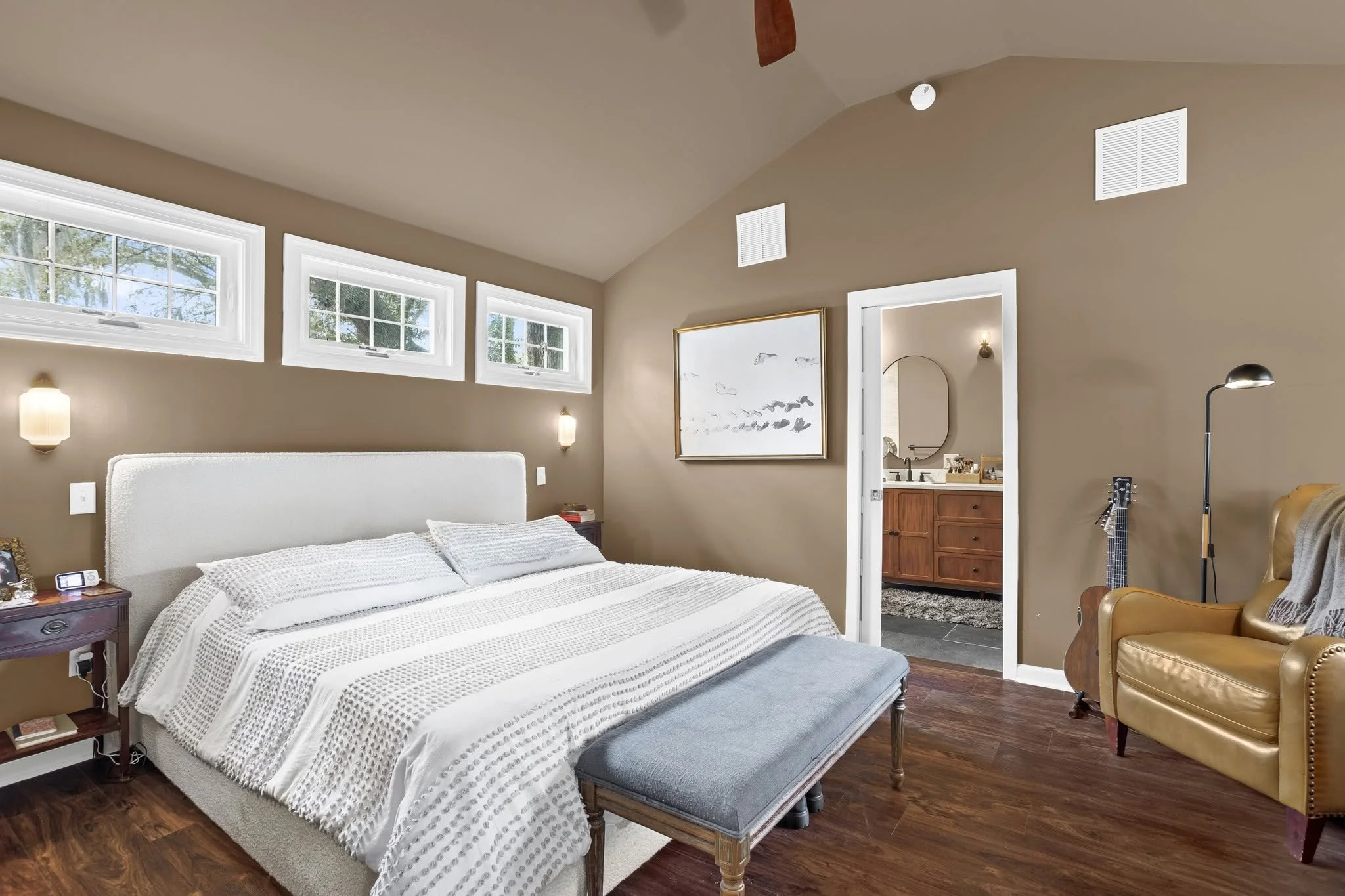 A bedroom with a white bed, beige walls, three high horizontal windows, a small nightstand, a mirror, a yellow armchair, and a doorway leading to a bathroom.