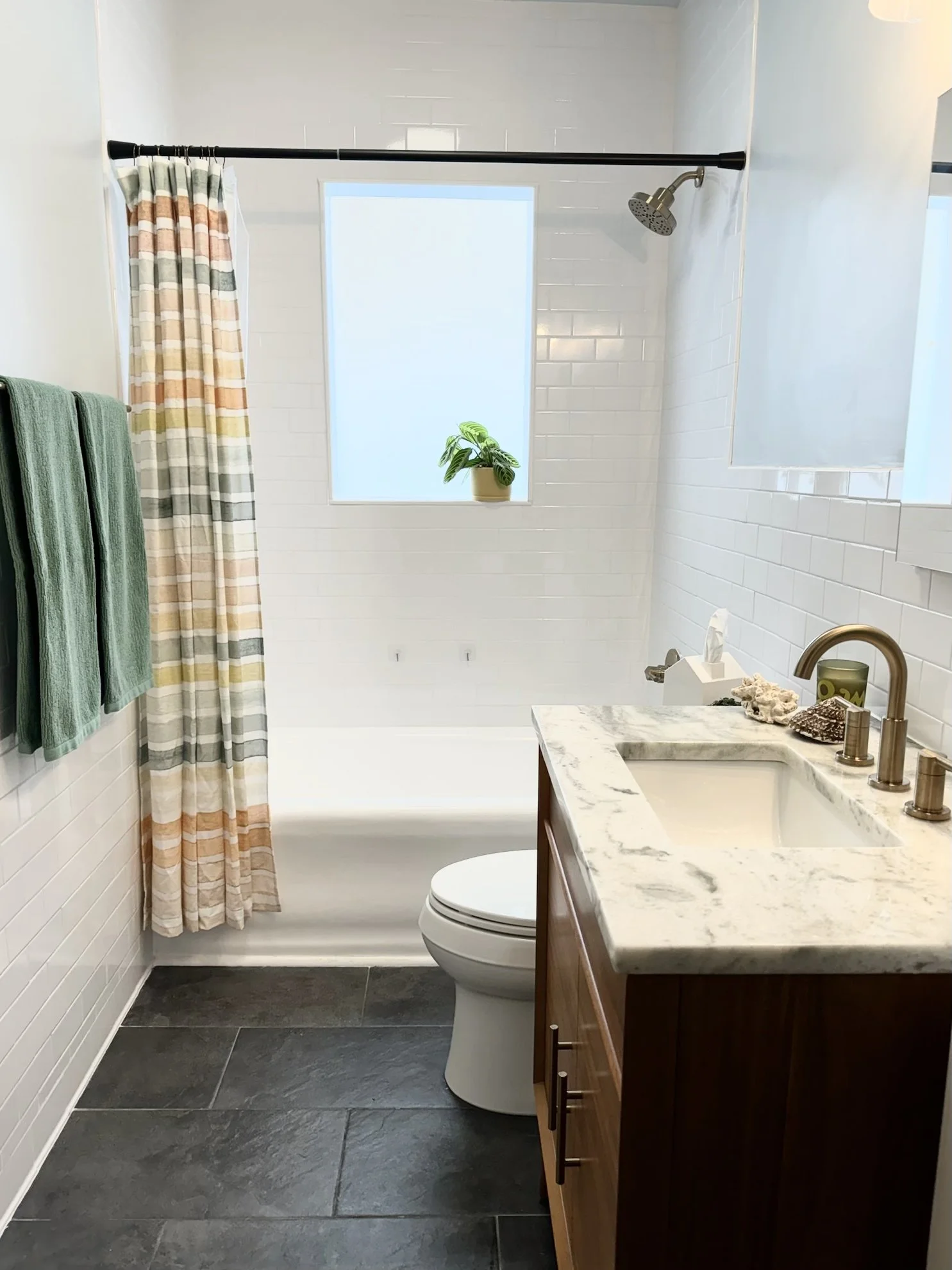 A modern bathroom with a vanity, marble countertop, and sink on the right, a toilet in the center, a white shiplap wall, a frosted window with a potted plant, green towels on a rack, a colorful curtain with stripes, and dark tiled floor.