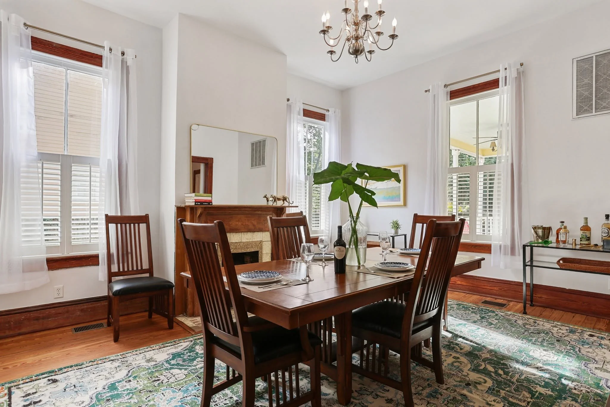 A dining room with a wooden table set with plates, wine glasses, and a bottle of wine, surrounded by wooden chairs, with windows with white curtains and a chandelier overhead, and a fireplace with a mirror and books in the background.