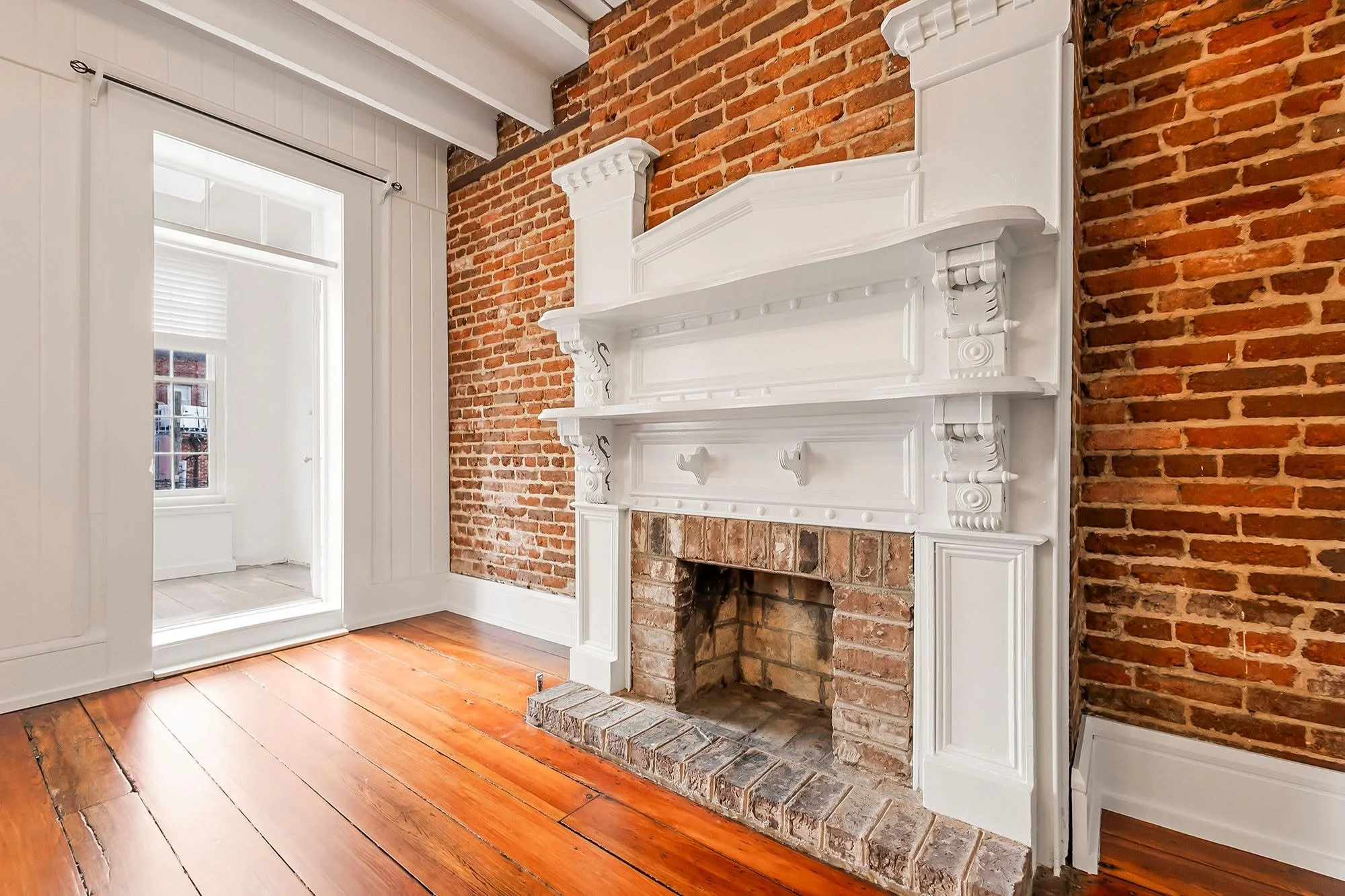 Interior of a room with a brick fireplace, white mantel, and wood flooring. A window with white trim is visible on the left, with another room visible through a doorway, featuring white walls and a window with blinds.