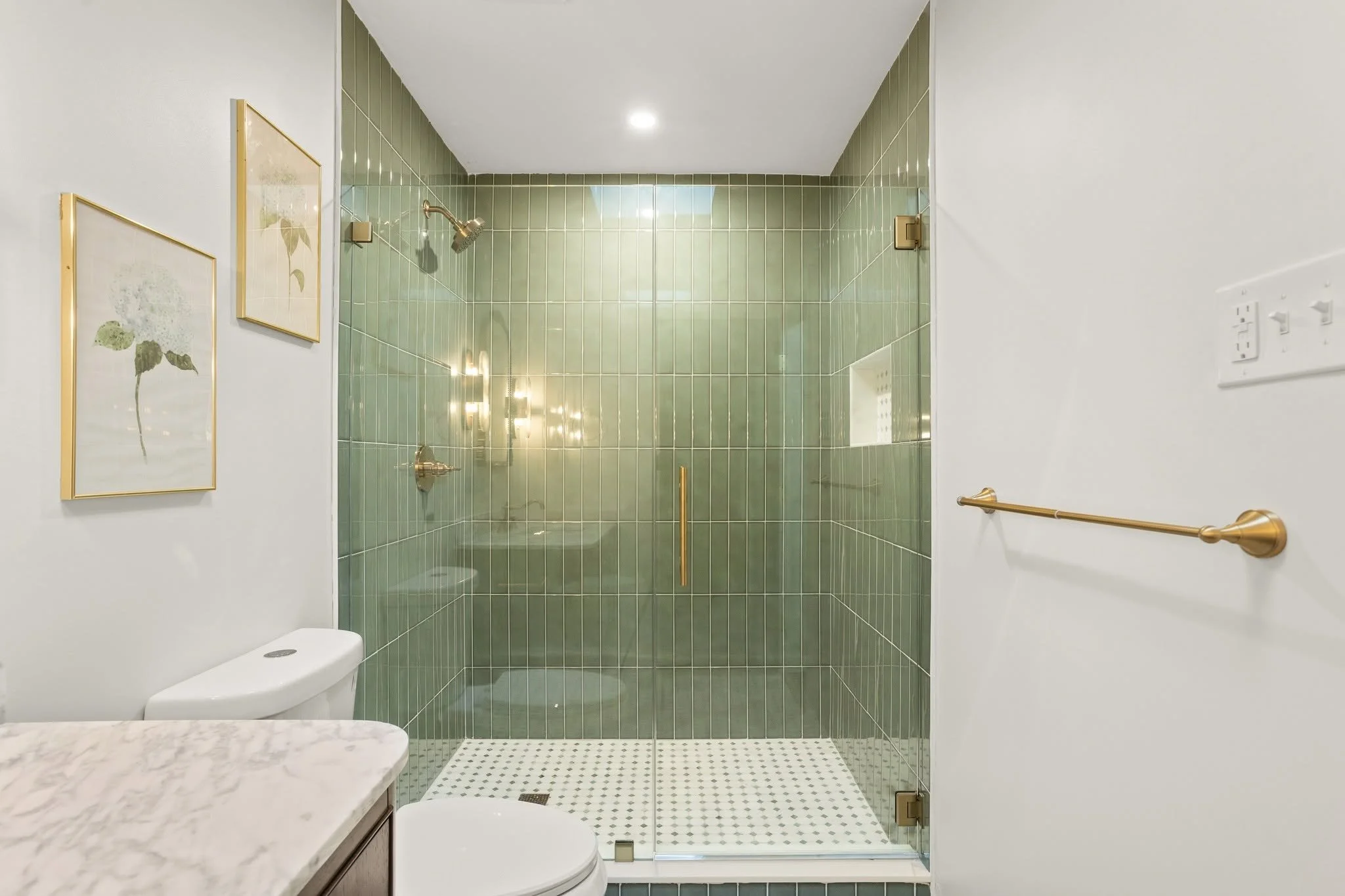 Bathroom with green tiled shower area, gold handle on glass door, white toilet, marble countertop, and framed botanical art on white wall.