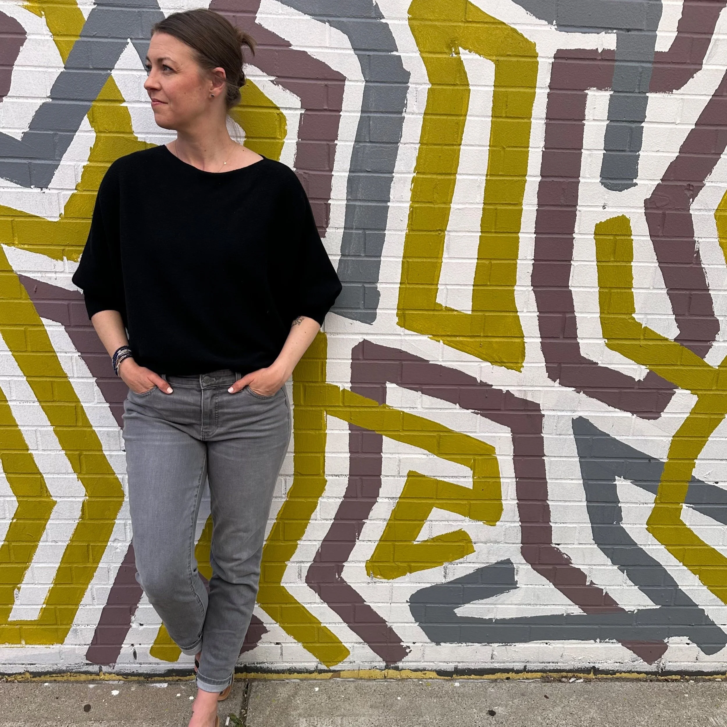 A woman standing in front of a colorful abstract mural on a brick wall, with only part of her body visible, wearing a black top and gray jeans.