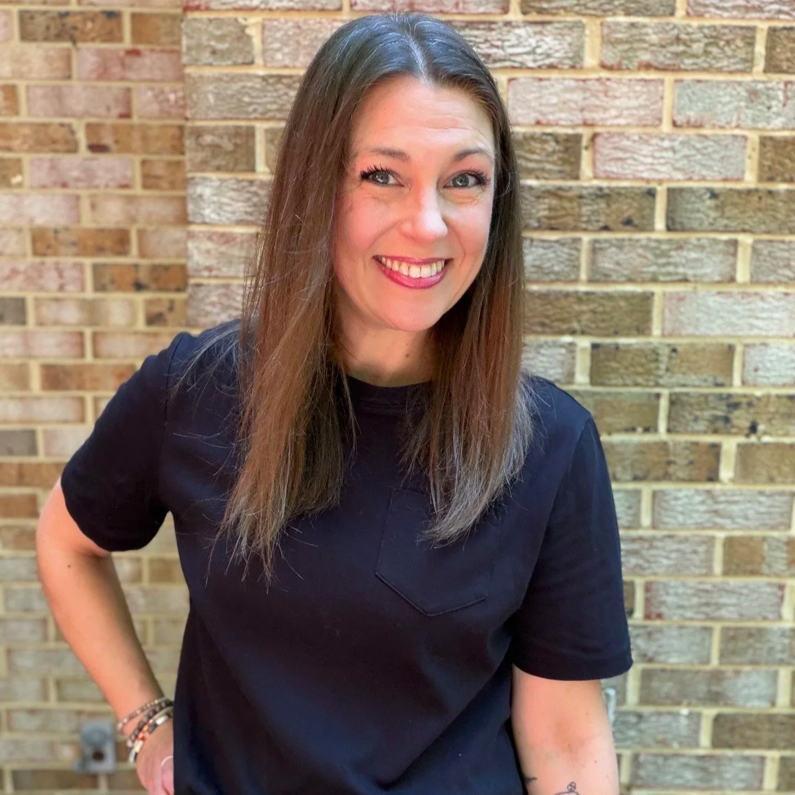 A smiling woman with long brown hair wearing a navy blue shirt standing in front of a brick wall.