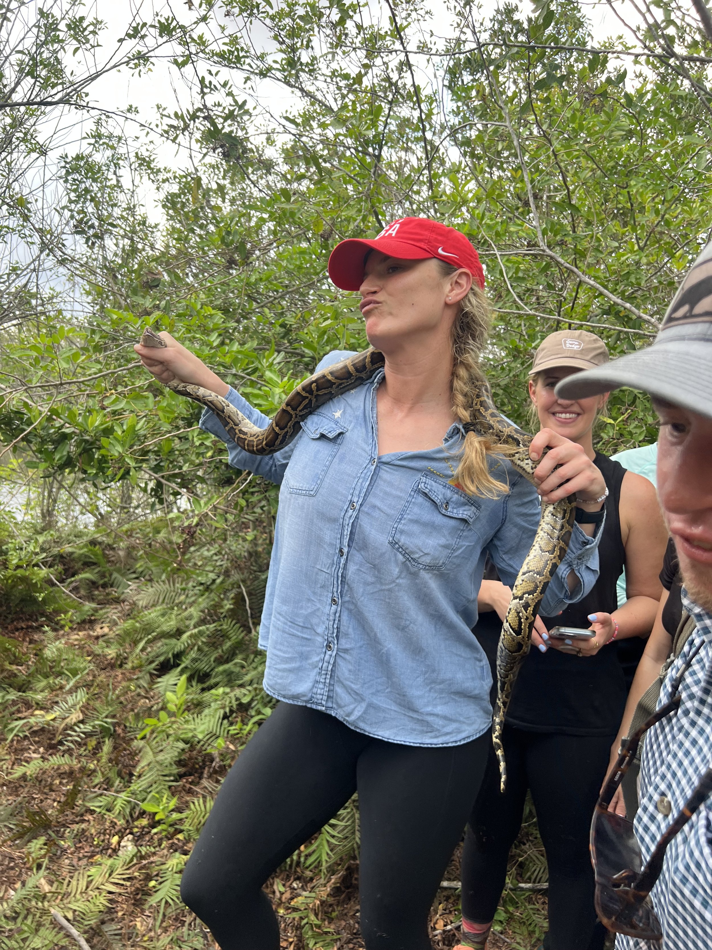 Woman in a red hat holding a large Burmese python across her shoulders outdoors