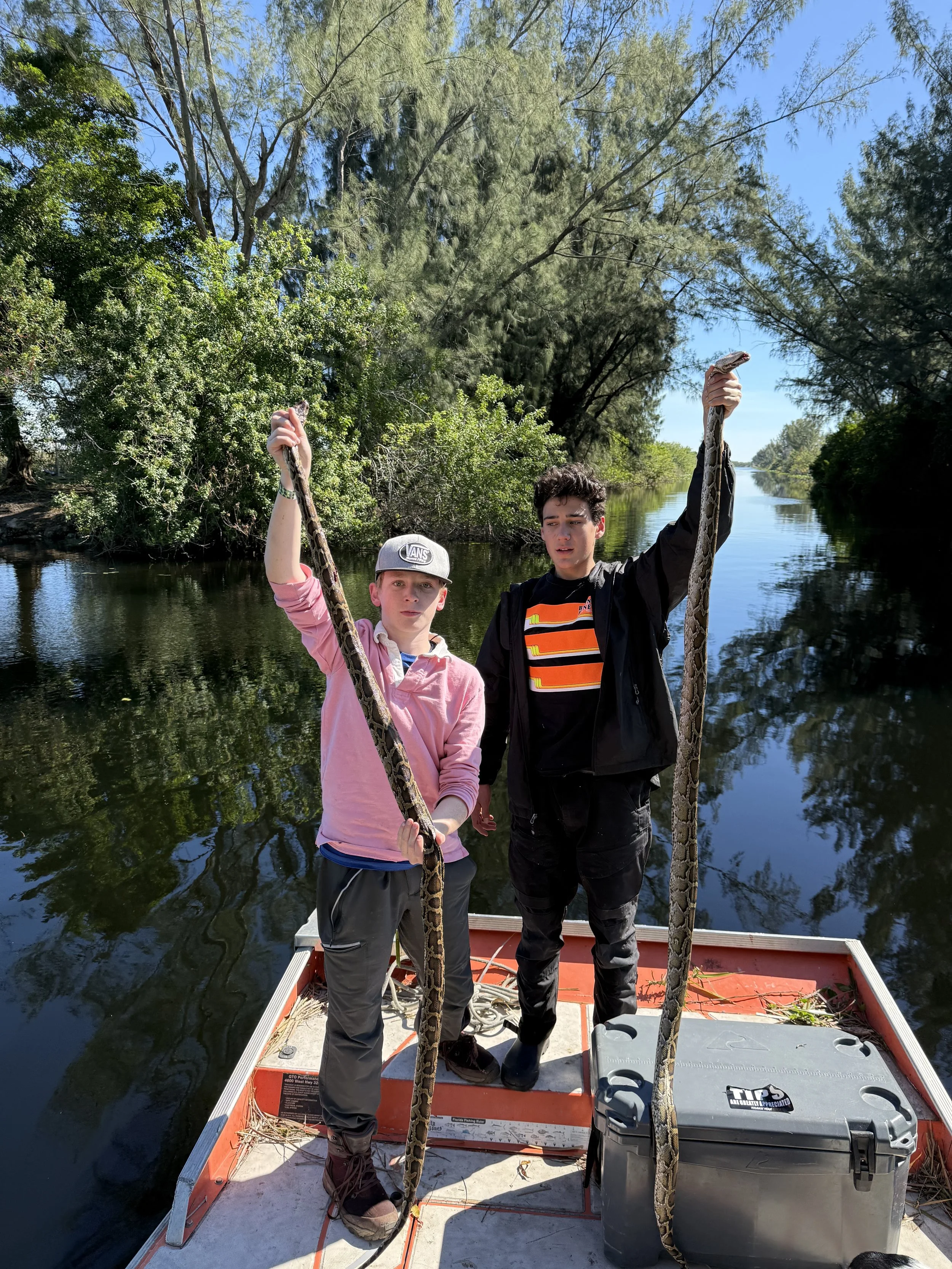 Two boys holding a large snake aboard an airboat on a tree-lined waterway