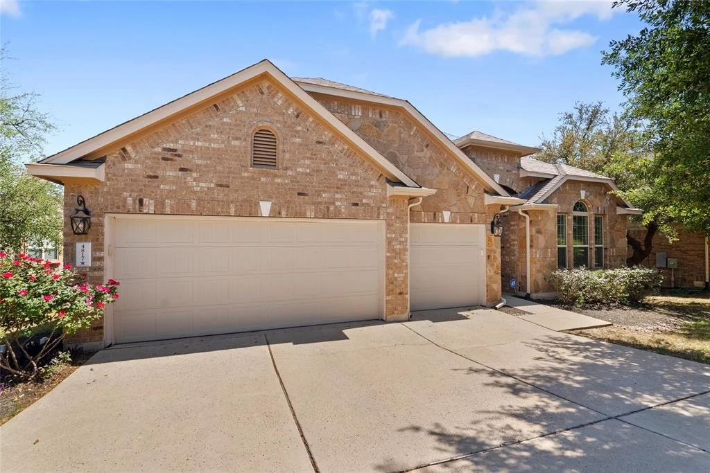 Front view of a brick house with a three-car garage, concrete driveway, and landscaped front yard with bushes and a flowering plant on the left side.