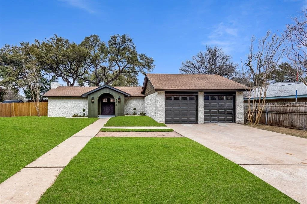 Front view of a house with white brick walls, green trim around the main entrance, a double garage with black doors, a concrete driveway, and a well-maintained lawn.