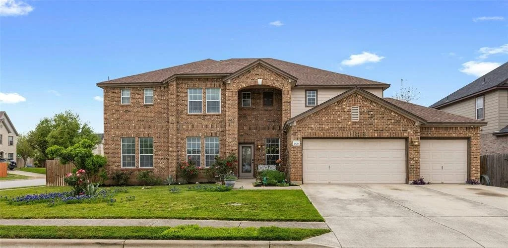Two-story brick house with a three-car garage and a well-maintained front yard under a partly cloudy sky.