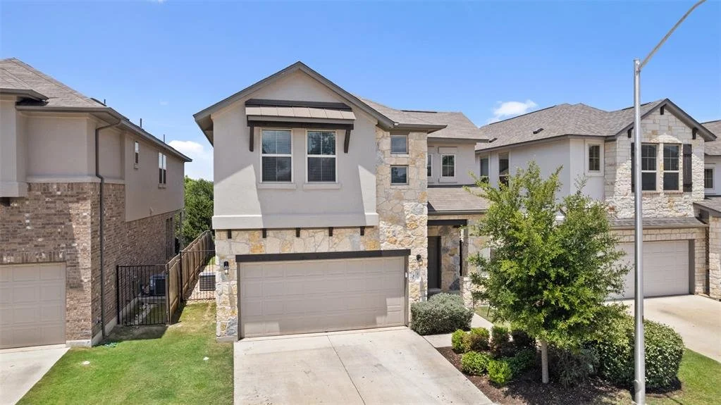 Modern two-story house with stone and stucco facade, attached garage, small front yard with shrubs and a tree, in a suburban neighborhood.