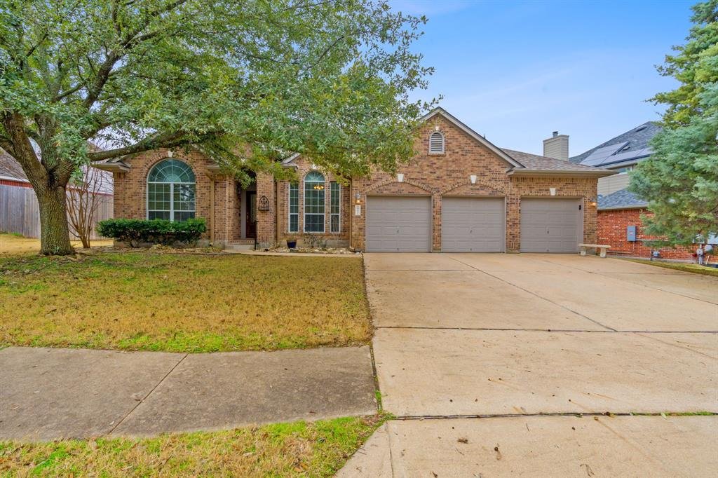 Front view of a brick house with a three-car garage, large front lawn, trees, and a concrete driveway.