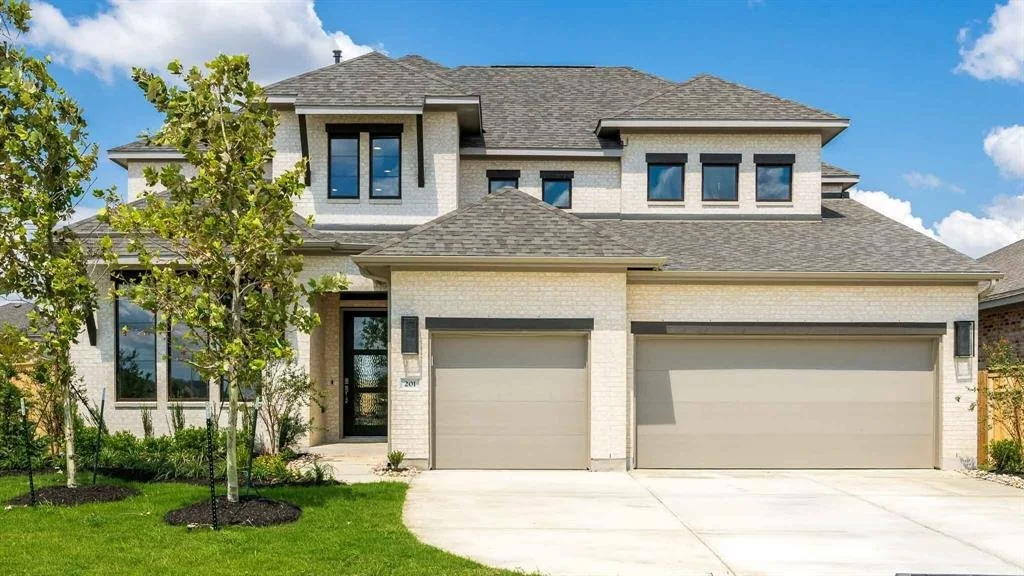 A modern two-story house with light-colored brick facade, gray roof, and three-car garage, surrounded by a well-maintained lawn and trees.