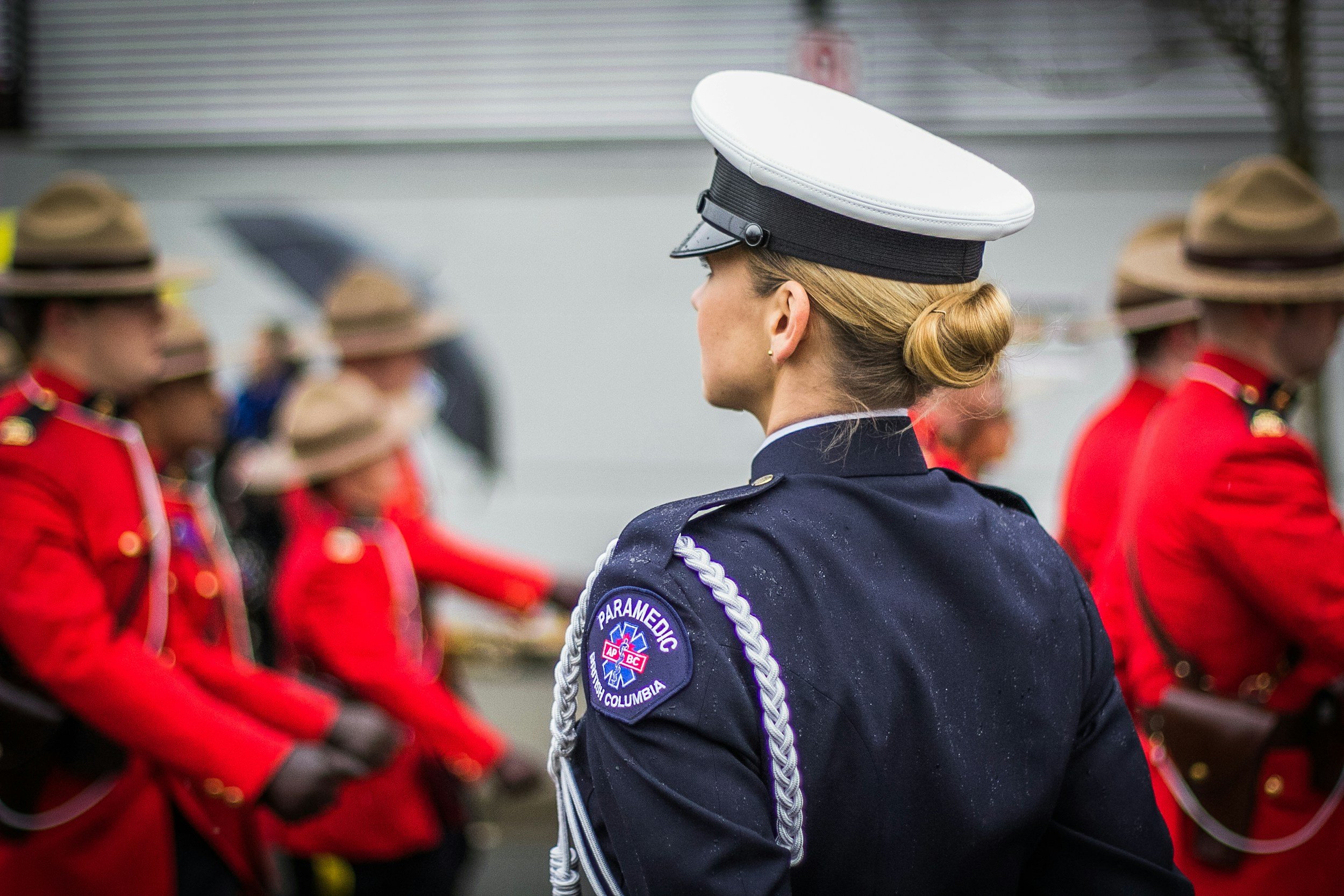 A female paramedic officer in uniform stands with her back to the camera, observing a line of people in red uniforms holding umbrellas. The scene appears to be outdoors in a formal setting.
