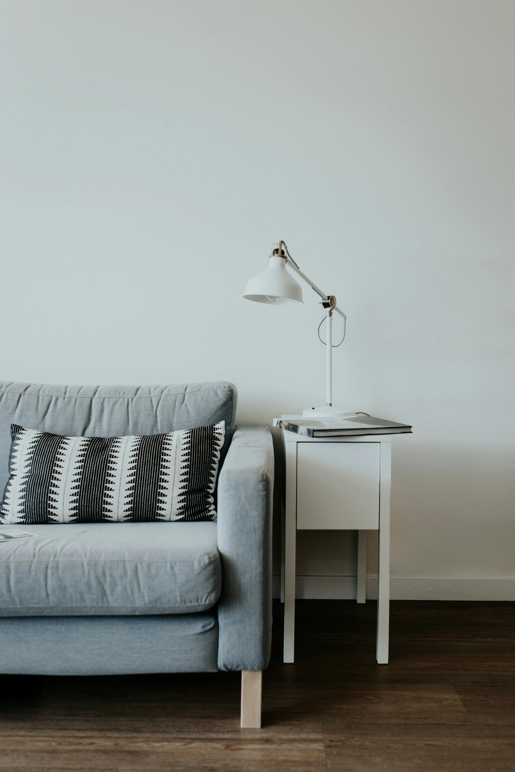 A light gray sofa with a black and white patterned pillow next to a small white side table. On the side table, there are magazines and a white adjustable desk lamp.