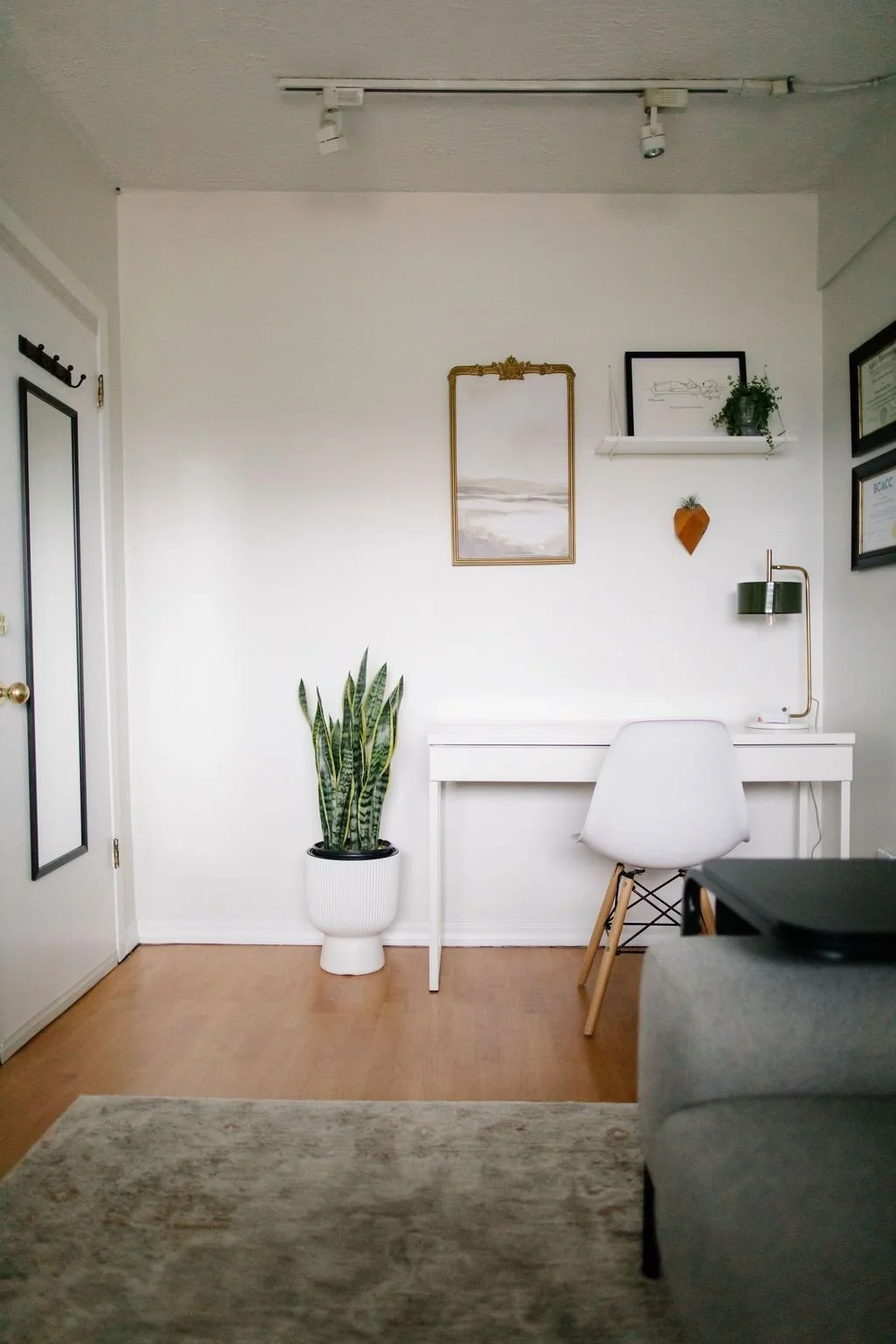 A minimalist office with a white desk and chair, a potted snake plant, framed artwork and certificates on the wall, a floating shelf with a plant, a wall-mounted lamp, and a small decorative wall hanging.