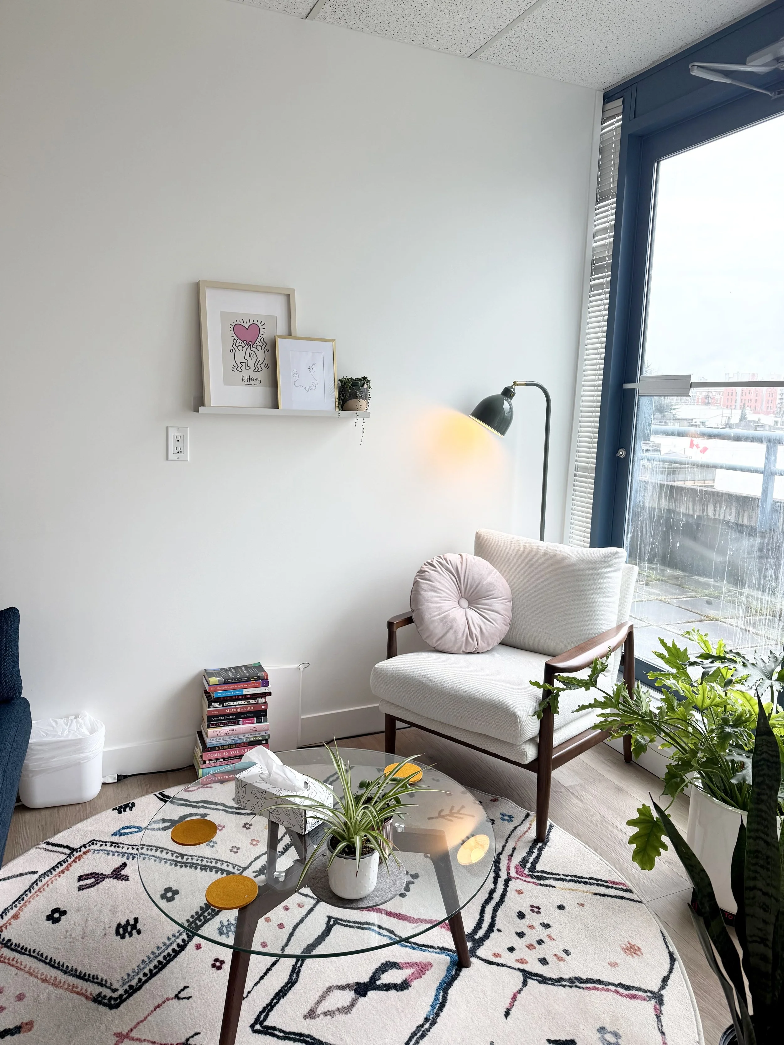 A cozy corner of a living room with a glass coffee table, a white armchair with a pink round pillow, a tall green plant, a floor lamp, and two picture frames on a small shelf on the wall. It is next to large windows with blinds, overlooking a cityscape. There's a stack of books on the floor, a small tissue box on the table, and a patterned area rug on the wooden floor.