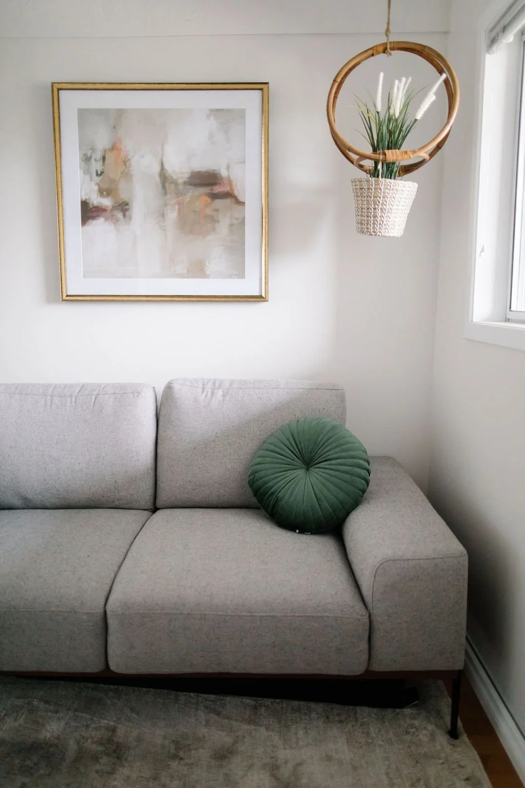 A beige sofa with a round, pleated green pillow on the right side. A framed abstract painting with soft, neutral tones hangs on the white wall above. To the right, a hanging planter with green plants is suspended from a circular wooden frame near a window.