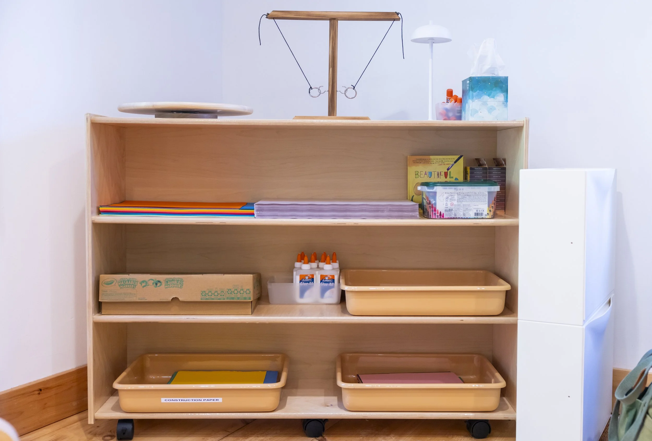 Wooden shelving unit on wheels containing art supplies, books, glue, paper, and storage boxes in a classroom or craft room.