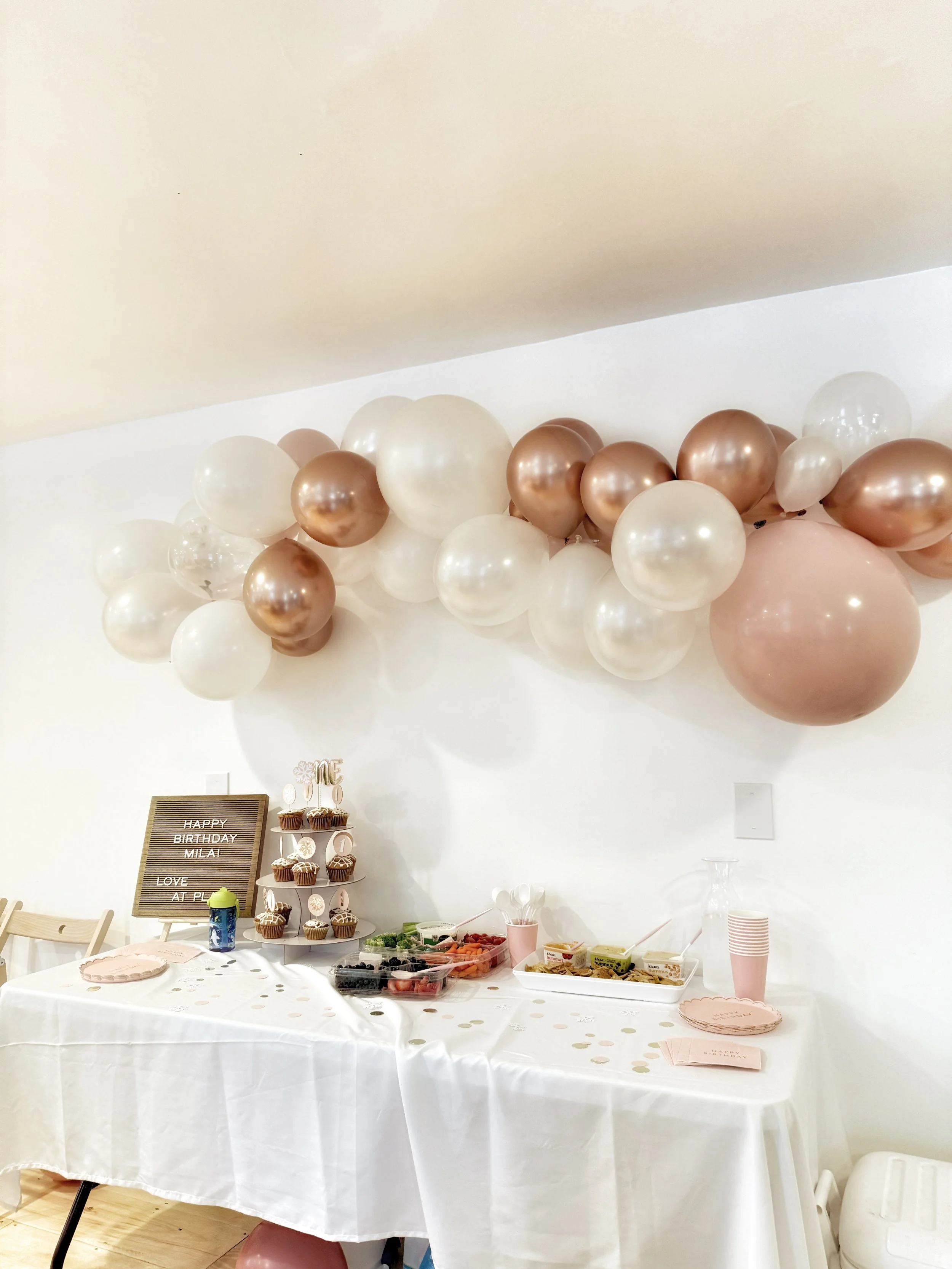 Birthday party setup with a white table, pink disposable cups, assorted food, and a balloon decoration on the wall made of white, metallic peach, and pearl-colored balloons.