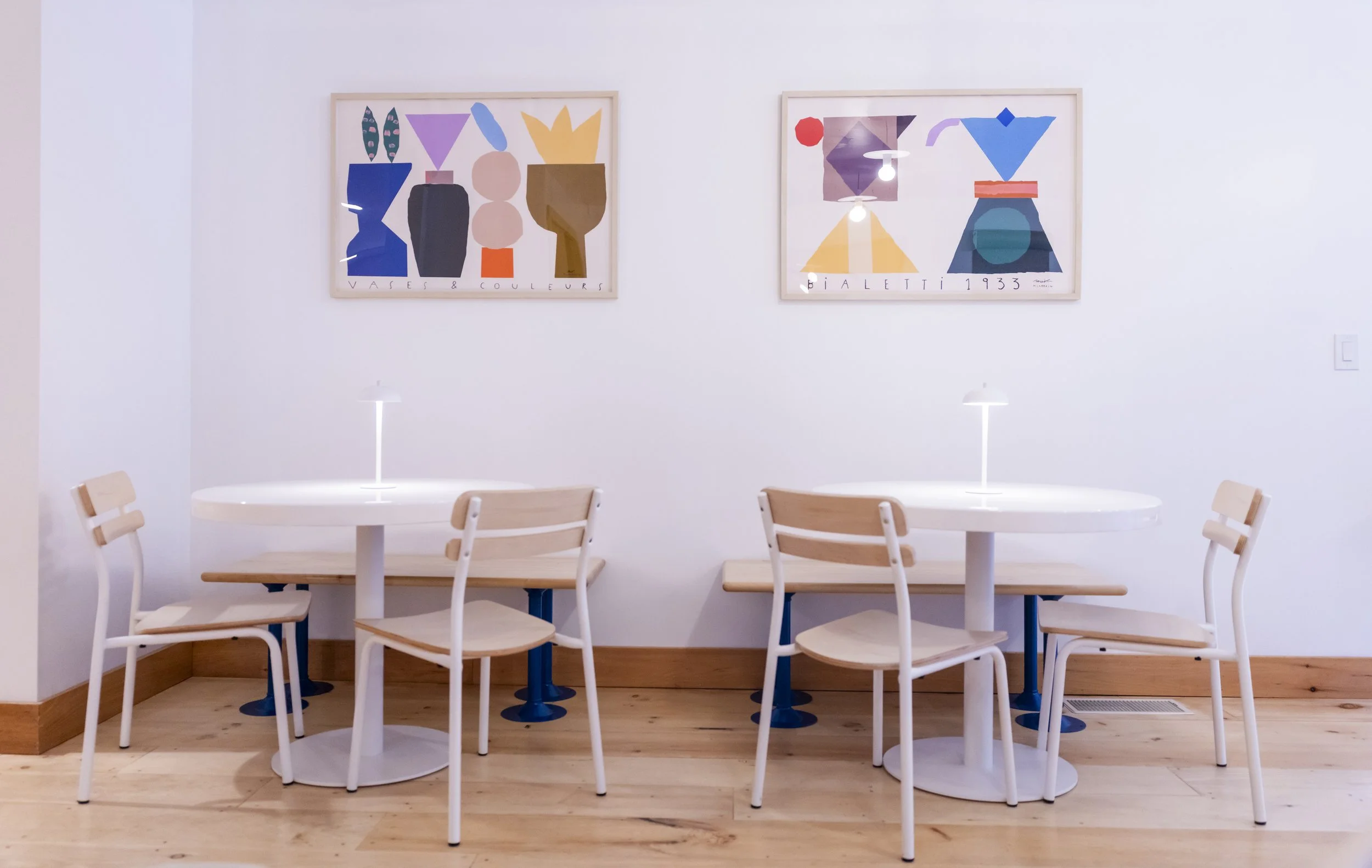 Empty dining area with two round white tables, four wooden chairs with white frames, and two framed abstract art pieces on a white wall.