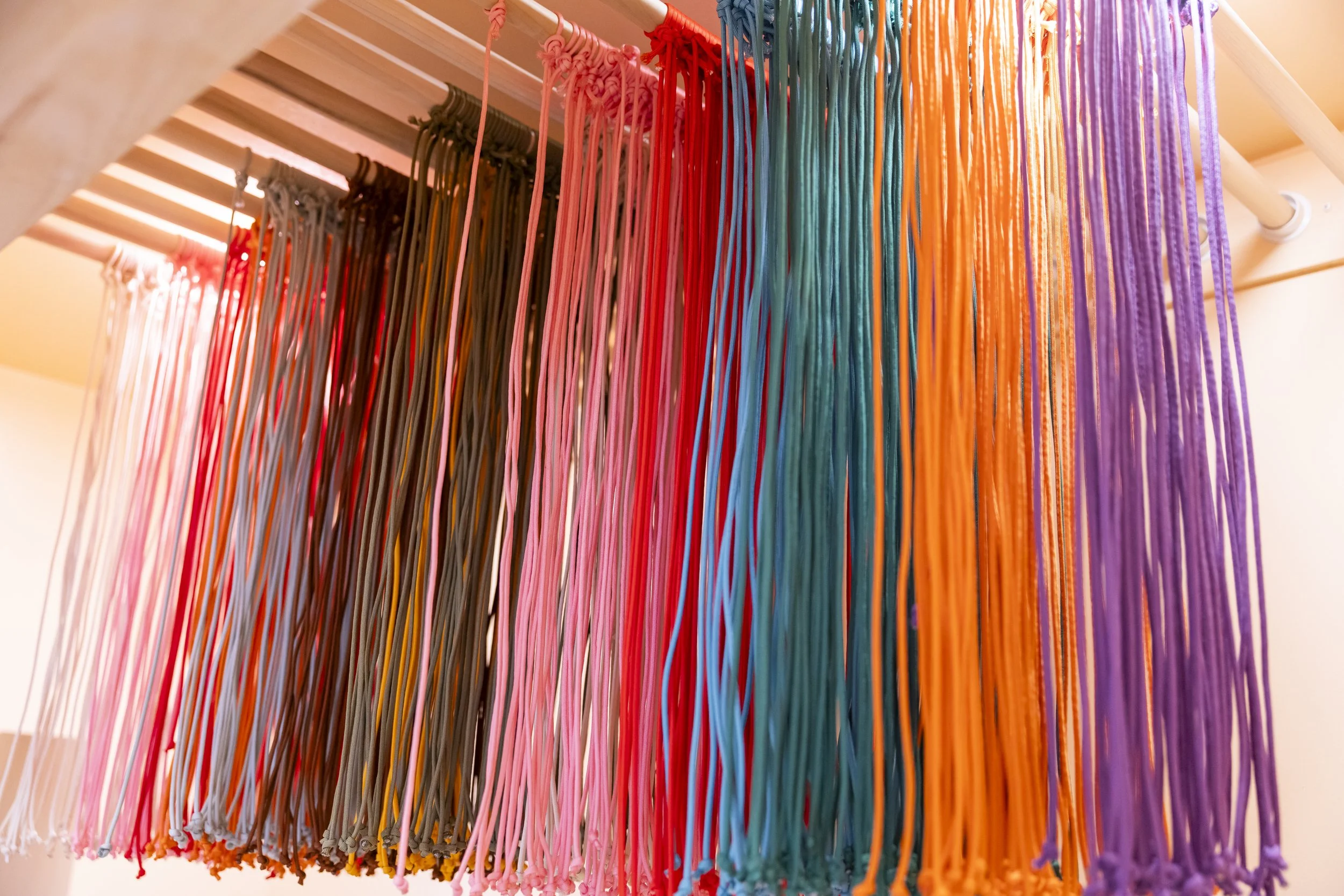 Colorful yarn strands hanging from a wooden rod, organized by color from dark to light to rainbow hues.