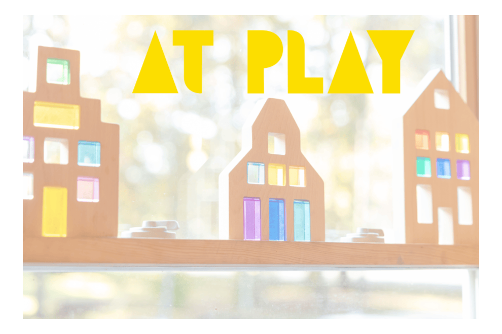 Colorful wooden building blocks arranged on a windowsill with the phrase 'AT PLAY' in bold yellow letters above.