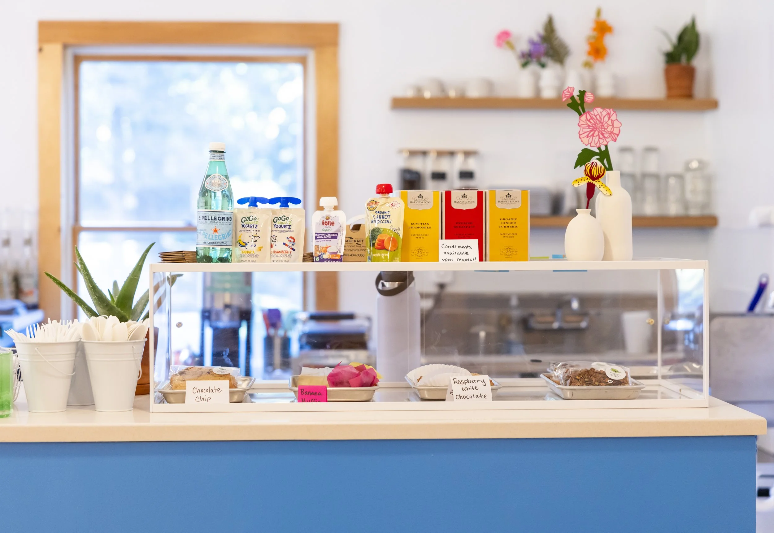 A display stand in a cafe or restaurant with bottled water, yogurt drinks, and boxes of tea, a potted aloe vera plant, and packaged snacks like chocolate chip cookies, banana muffins, and raspberry white chocolate muffins, in a bright, cozy setting w