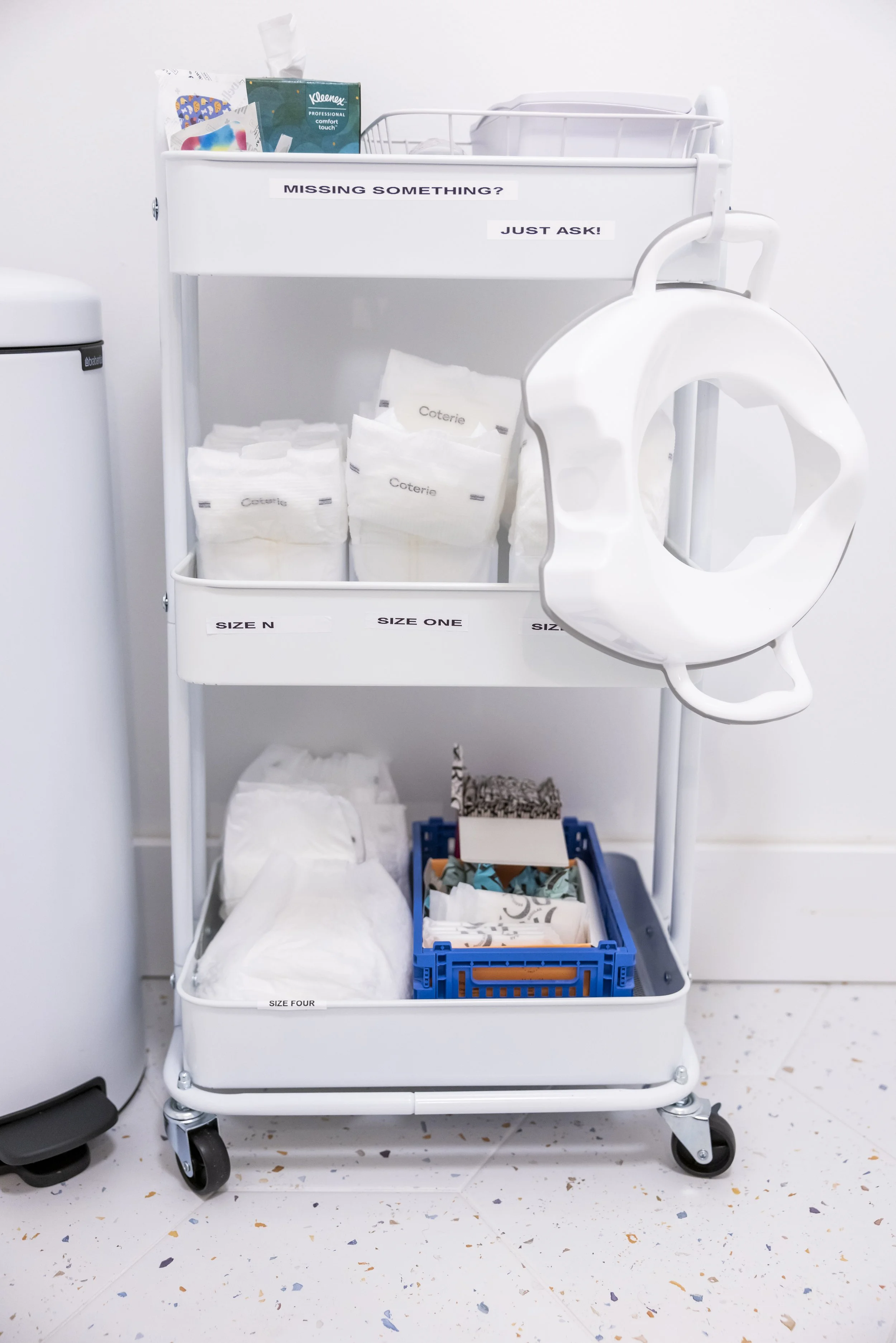 Medical supply cart with diapers and a tissue box, attached to a wall with a sign saying 'Missing Something? Just Ask!'. Diapers are organized by sizes labeled on the cart.