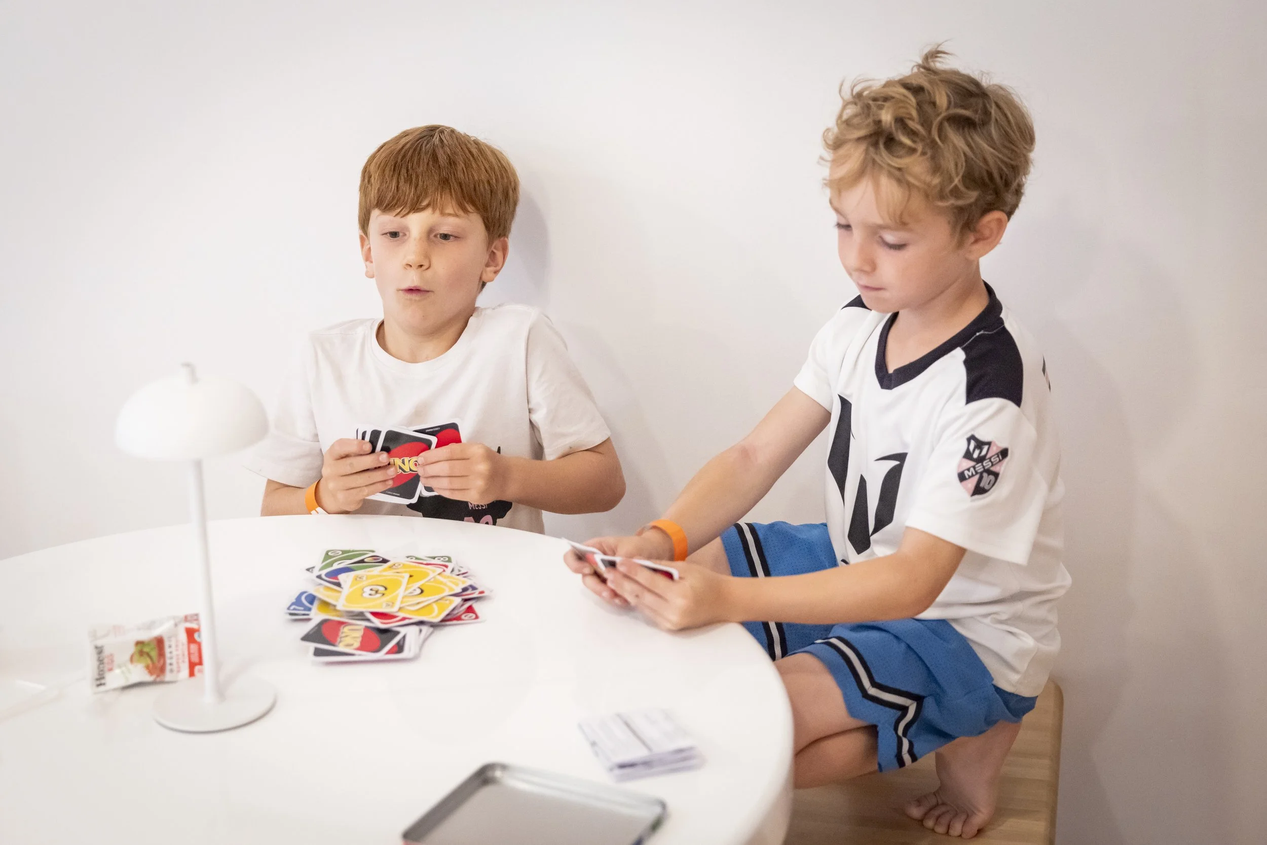 Two young boys sitting at a white table playing a card game, with a small lamp and a snack package on the table.