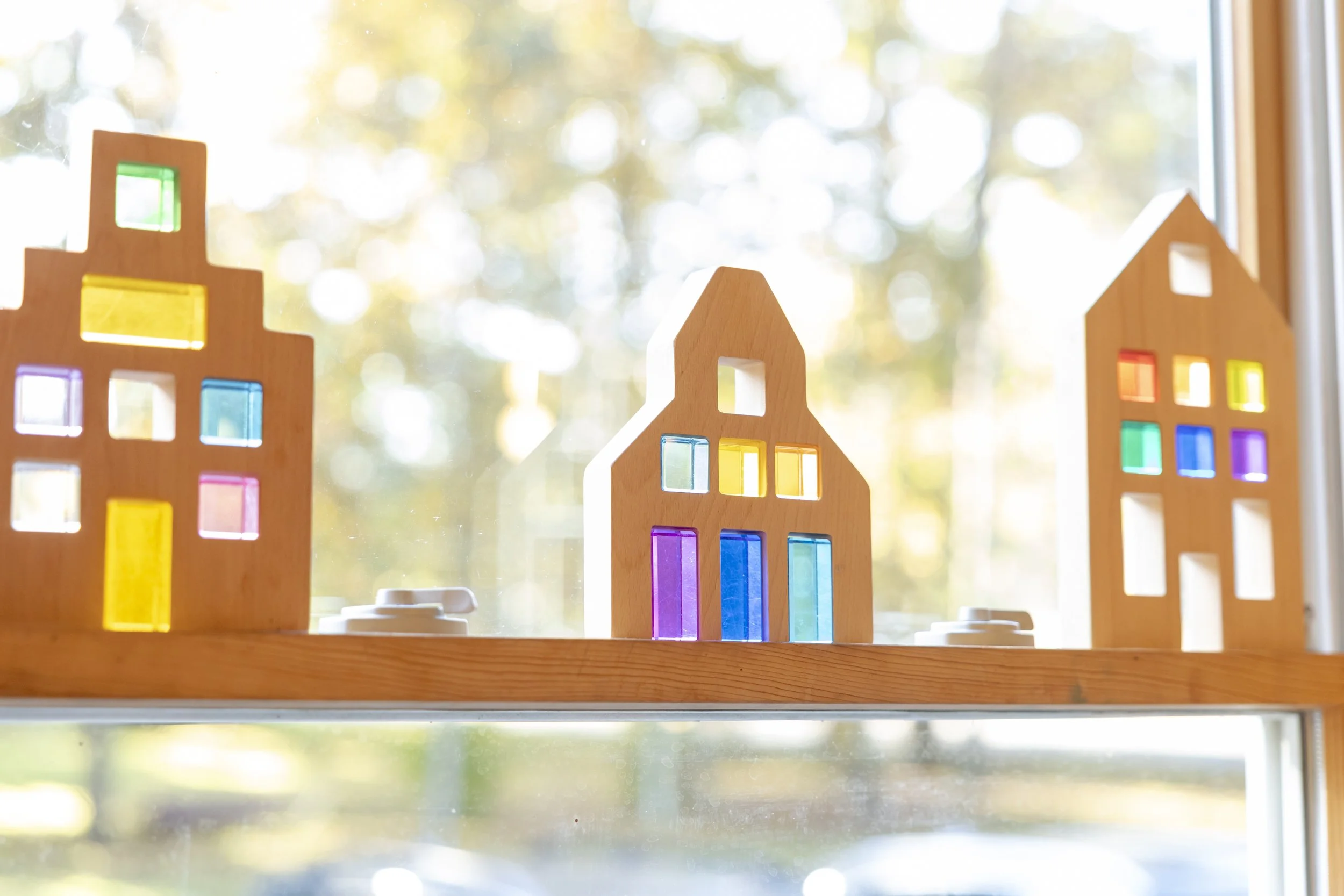 Wooden building toys with colorful stained glass windows placed on a window sill, blurred autumn trees in the background.
