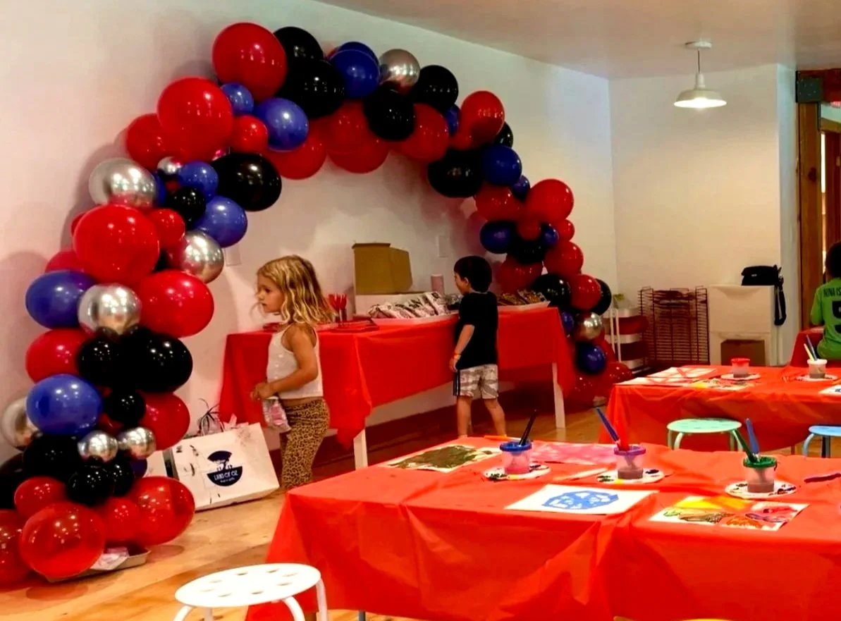 Children at a party with colorful balloons forming an arch, decorated tables with supplies, and kids engaging in activities.