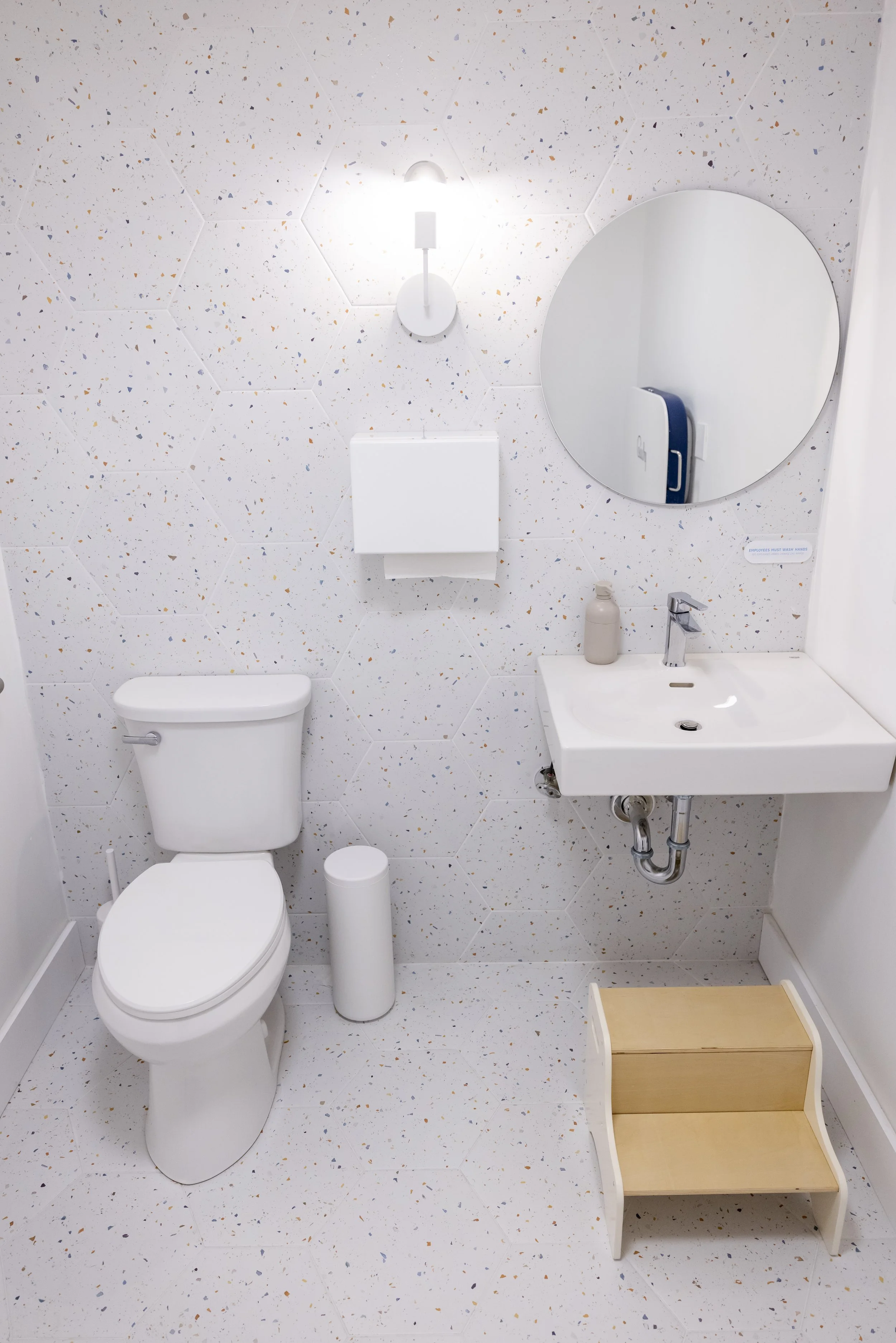 A small white bathroom with a toilet, a wall-mounted sink, a round mirror, and a wood-framed step stool. There is a wall light above the mirror and a soap dispenser on the sink.