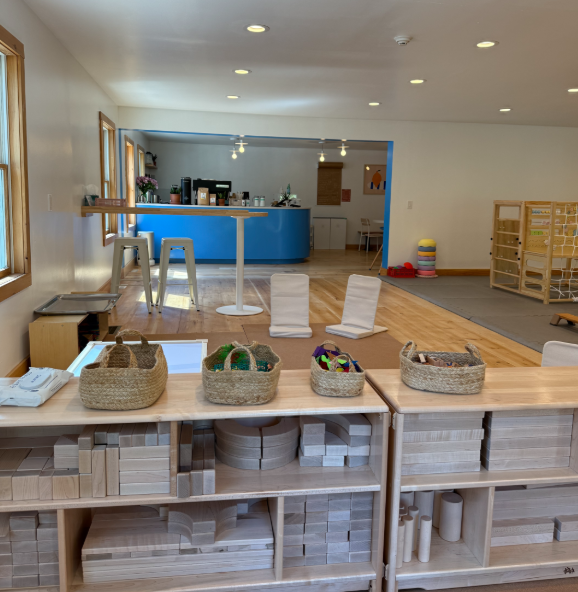 Childcare or playroom area with baskets of toys on a shelf, a countertop with a telephone, and a view into a larger room with chairs, a blue counter, and children's toys and décor.