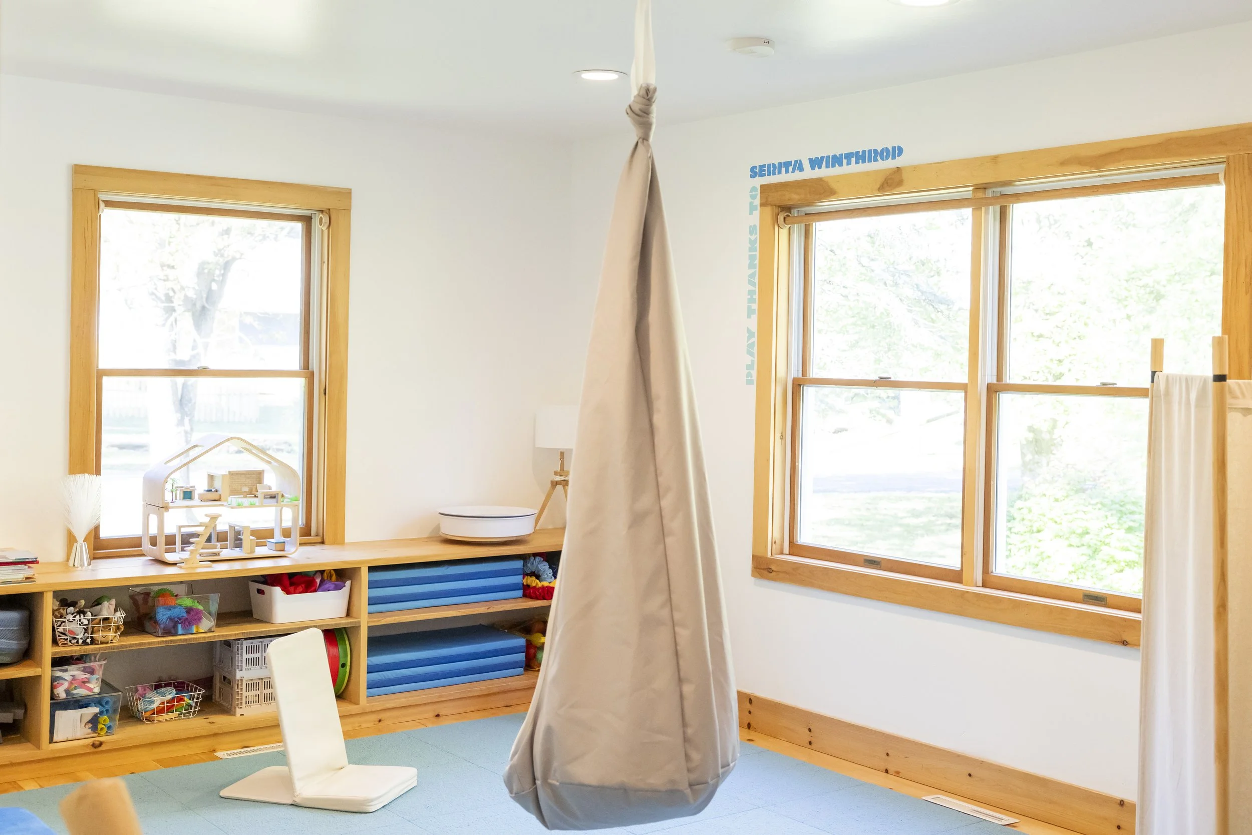 Child's playroom with wooden framed windows, a hanging fabric swing, a low wooden shelf with toys, and playmat on the floor.
