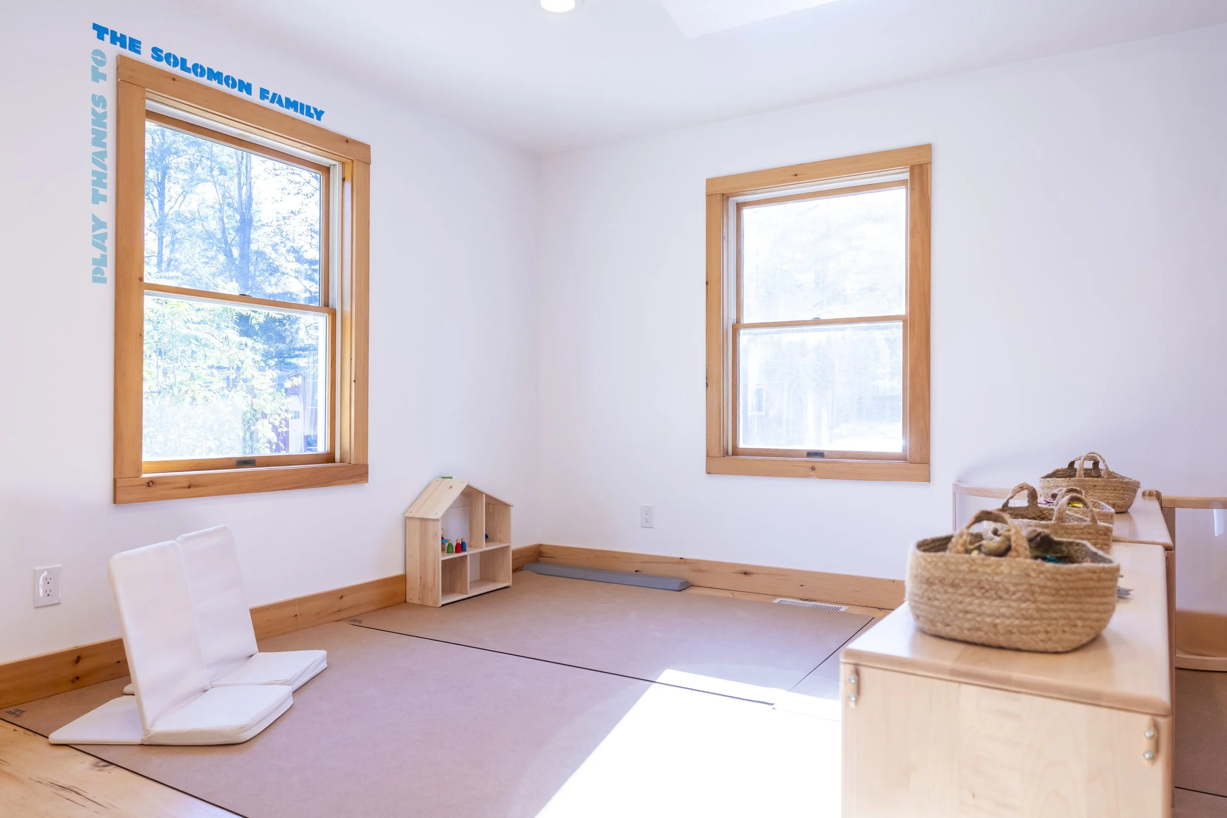 A bright, minimalist children's playroom with two large windows framed in natural wood. The room has a beige carpeted floor, and a small wooden house shelf with toys and figurines. There are several woven baskets on a wooden counter, a white foam puz