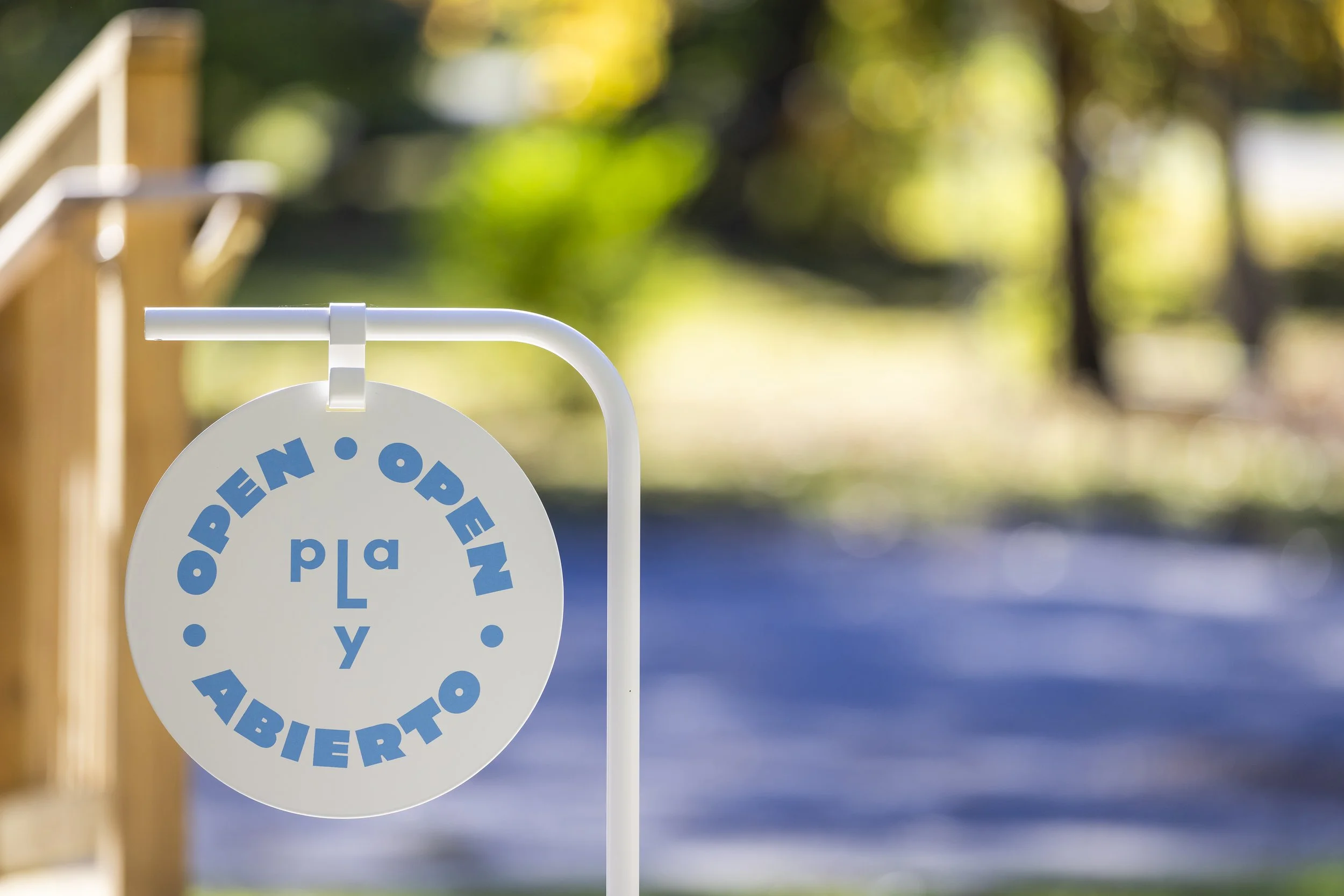 A sign hanging on a white stand outdoors with the words "Open" and "Abierto" in blue, and the word "Play" in the center.