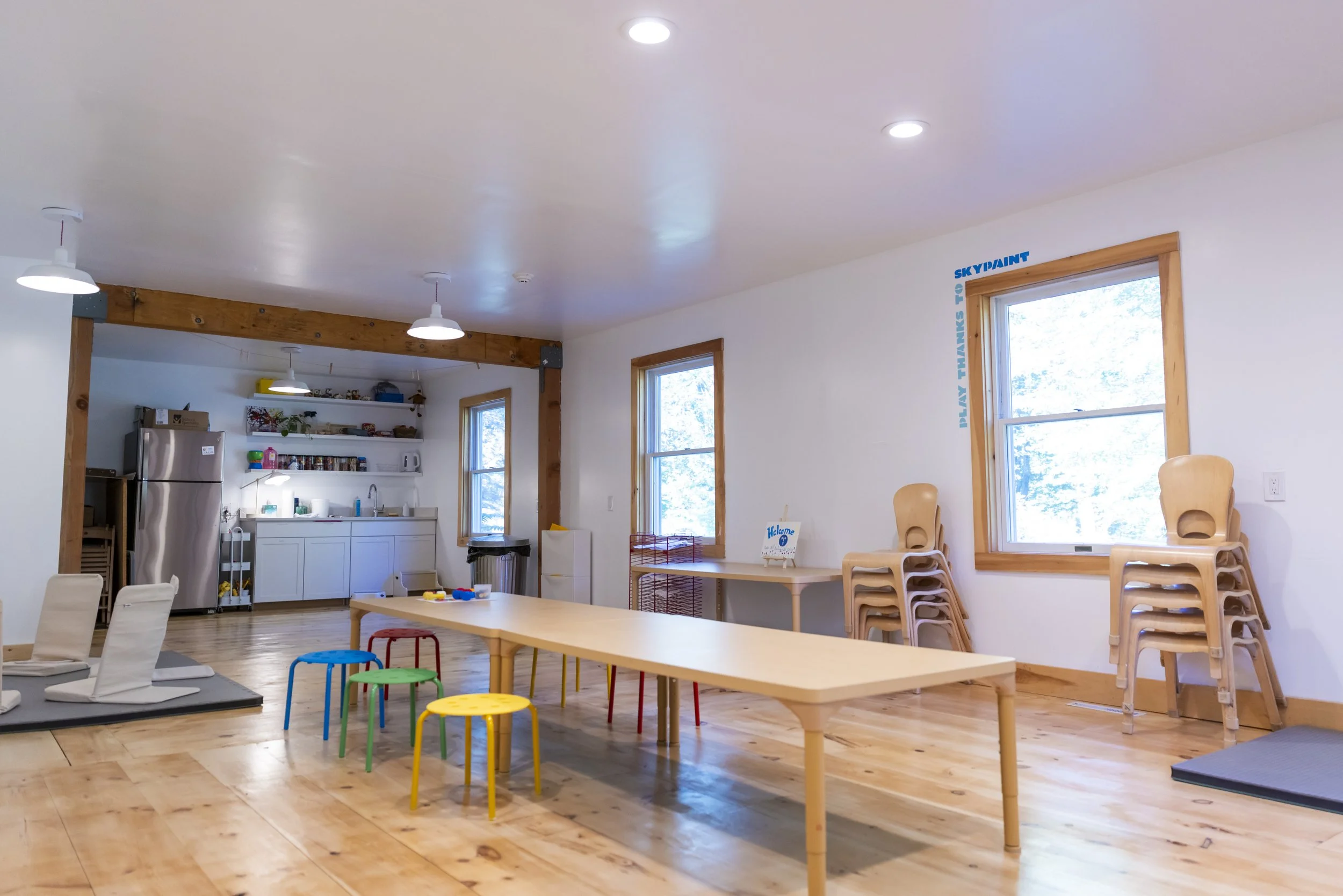 Bright and spacious daycare room with wooden floors, white walls, and large windows. There are small colorful chairs around a long wooden table, stacked wooden chairs near the windows, and a play area with a mat and toys. The kitchenette area include