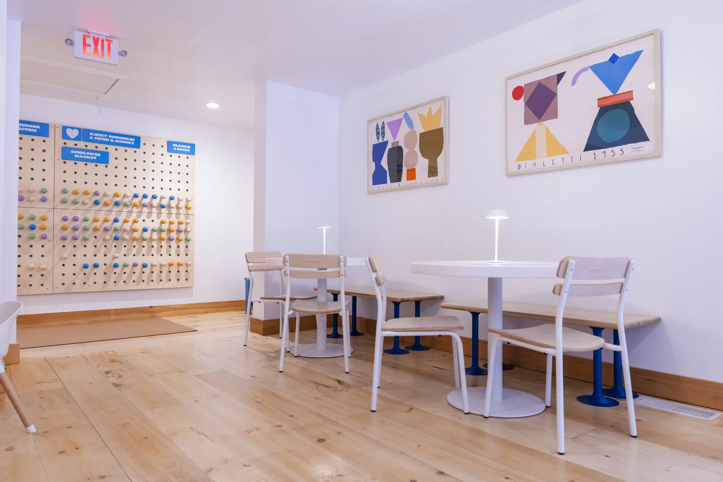 Empty dining area with white tables and chairs, colorful abstract artwork on the walls, a wooden pegboard with colorful hooks, and an exit sign on the ceiling.