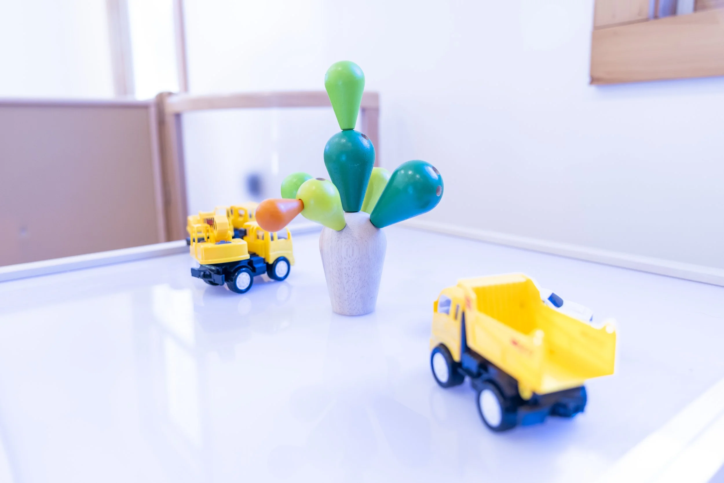 Decorative wooden vase with colorful wooden pegs resembling flowers, with two yellow toy dump trucks on a white table, in a room with white walls and wood accents.