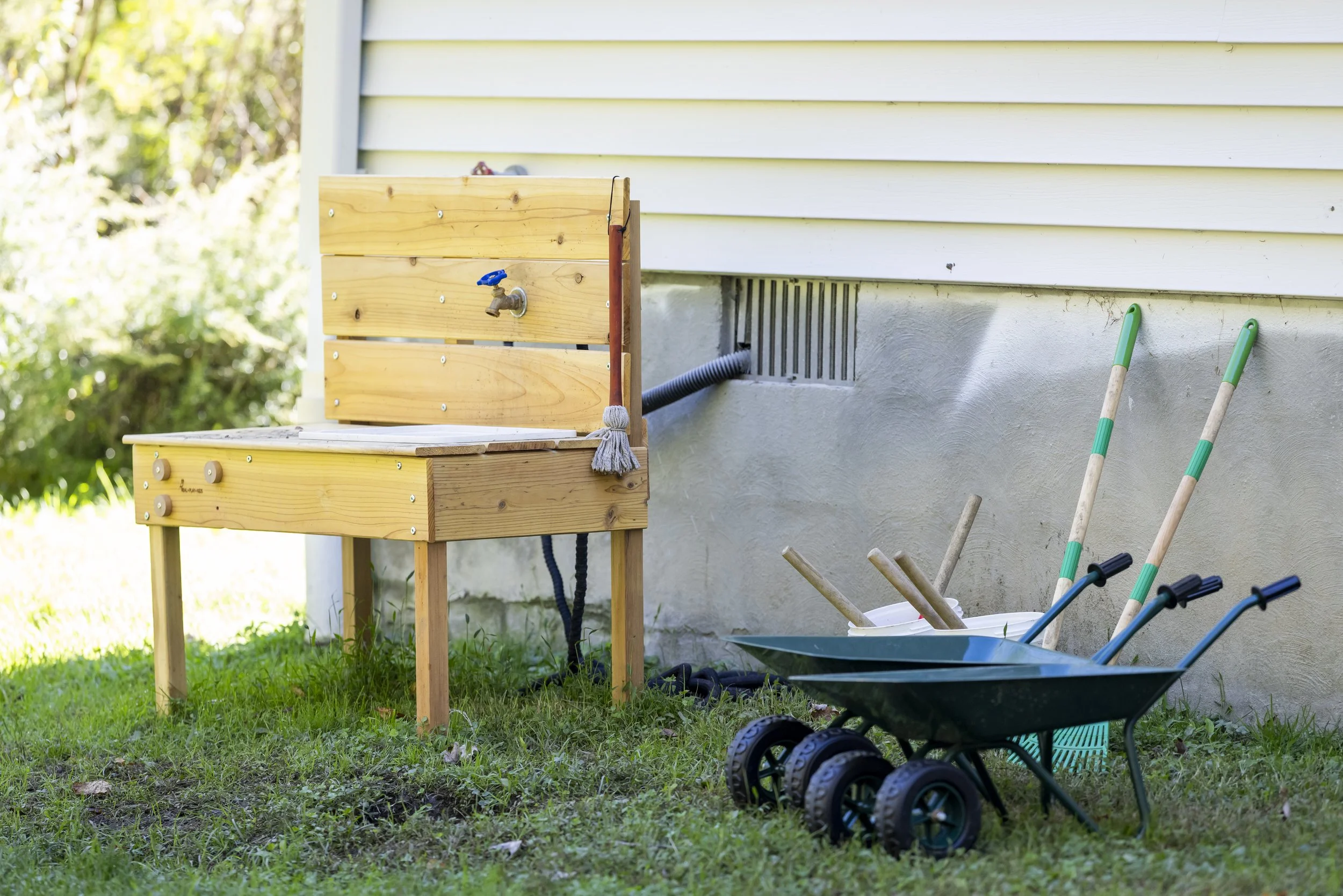Children's outdoor play area with a wooden play kitchen sink, a plastic wheelbarrow filled with gardening tools, and rakes leaning against a house exterior wall.