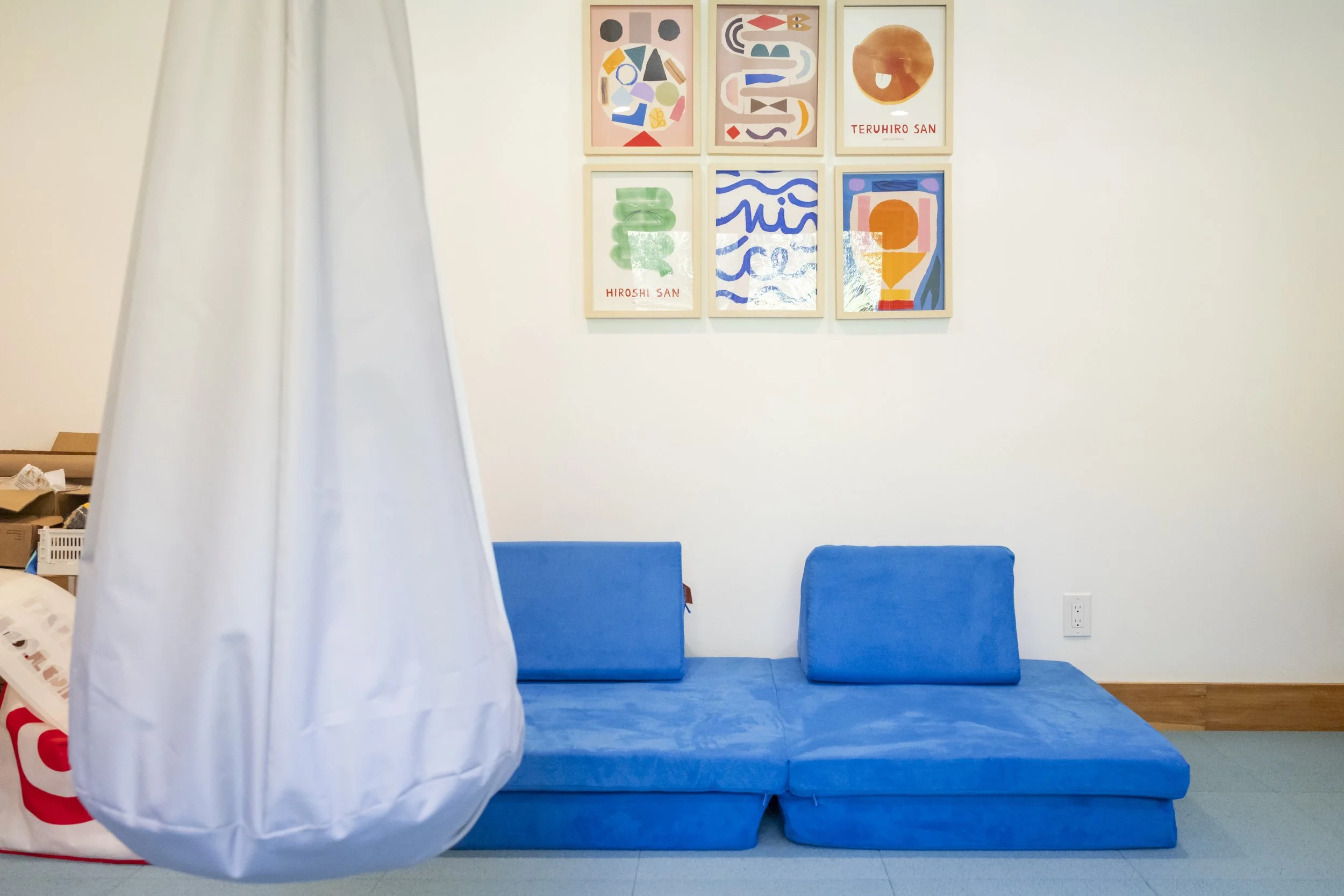 A blue cushioned seating area with two backrest cushions on a light green floor. To the left, a hanging white fabric is partially visible. The white wall behind features six colorful abstract art pieces in frames, with some including the names Hirosh