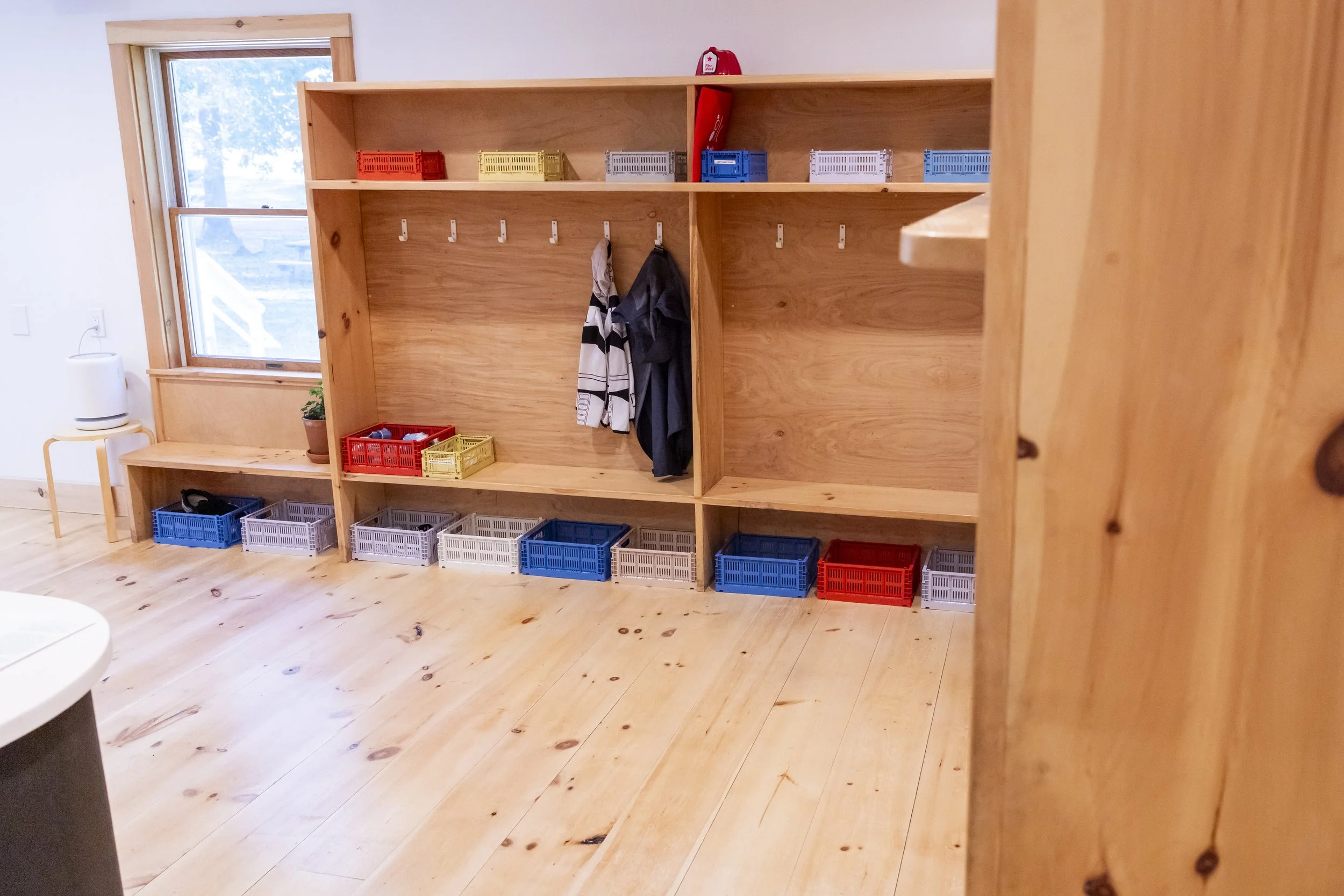 Empty wooden cubby storage with hooks, small plastic baskets, and a few coats hanging, near a window in a room with wooden floors.