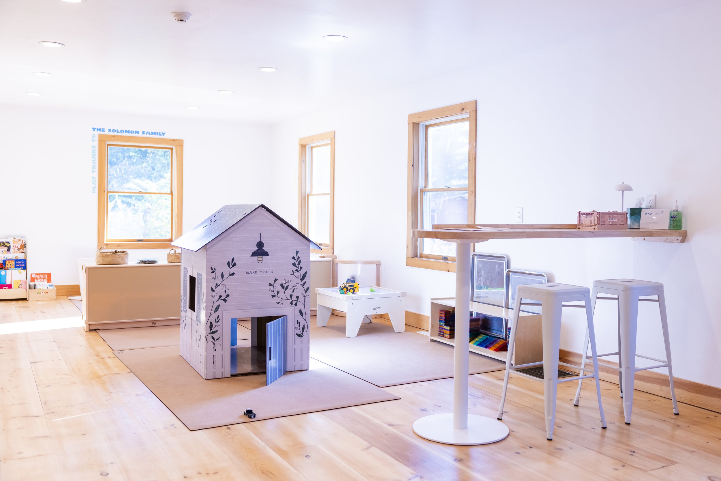 Bright children's playroom with wooden floors, three windows with wooden frames, and white walls. Features a playhouse with a gray roof and painted black decorations, a white table with colorful toys, a storage bench, and small shelves with books and