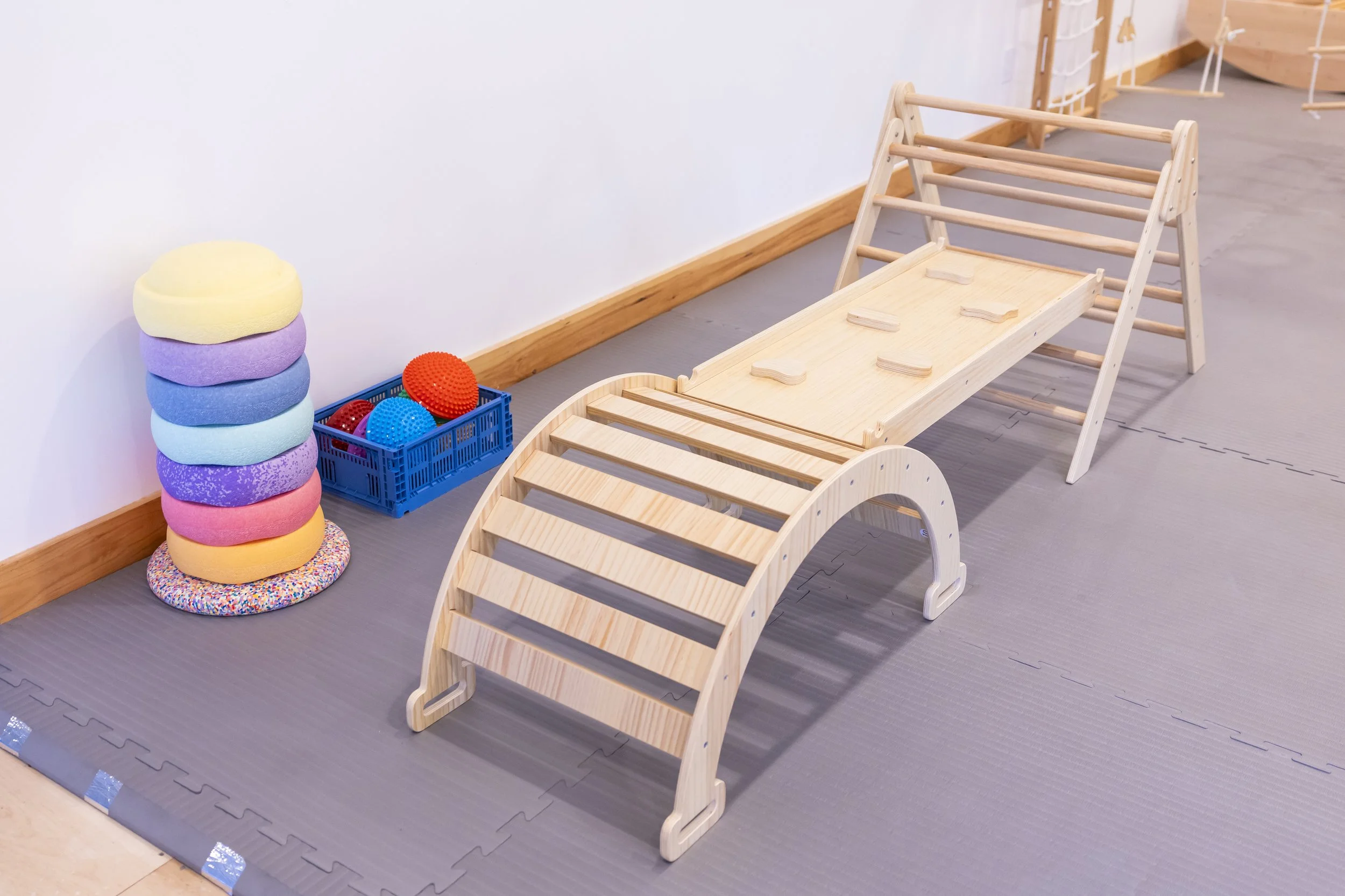 Wooden indoor climbing structure with ladder and climbing ramp, plush colorful cushions stacked in a corner, and a blue crate containing textured balls on a padded gray floor.