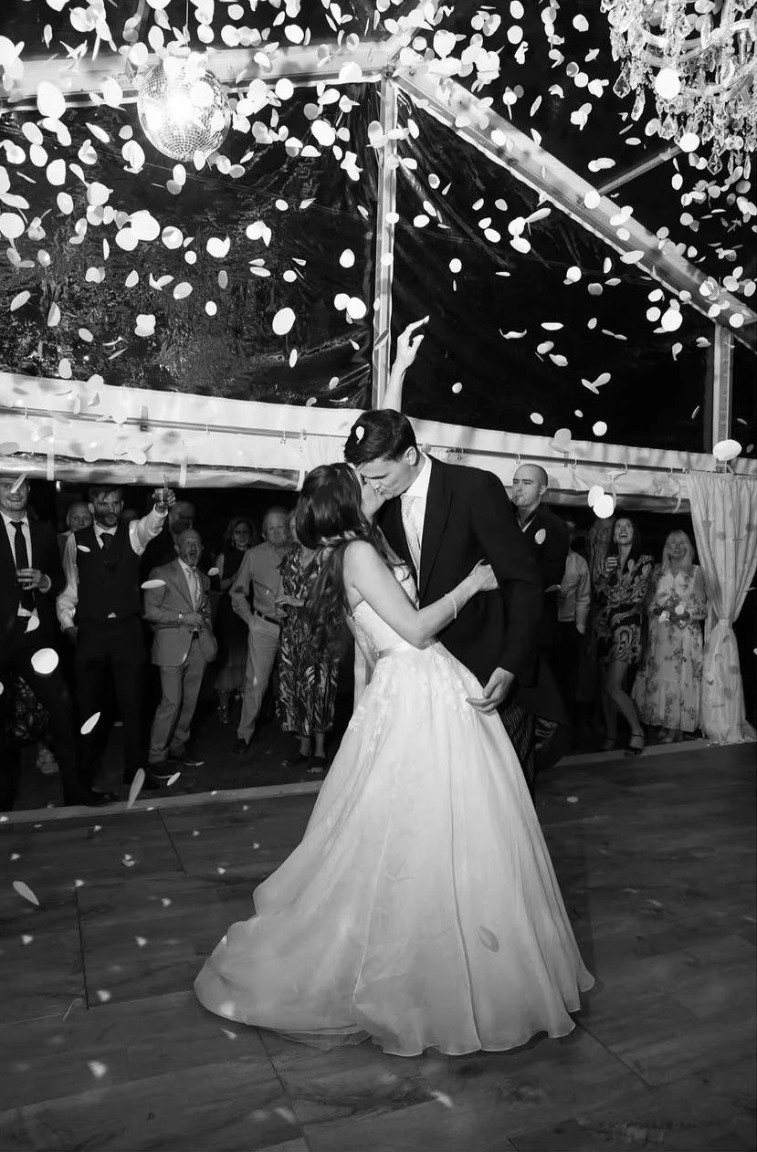 A couple in wedding attire dance closely at a wedding reception while guests surround them. Confetti falls from above, and a disco ball hangs from the ceiling.