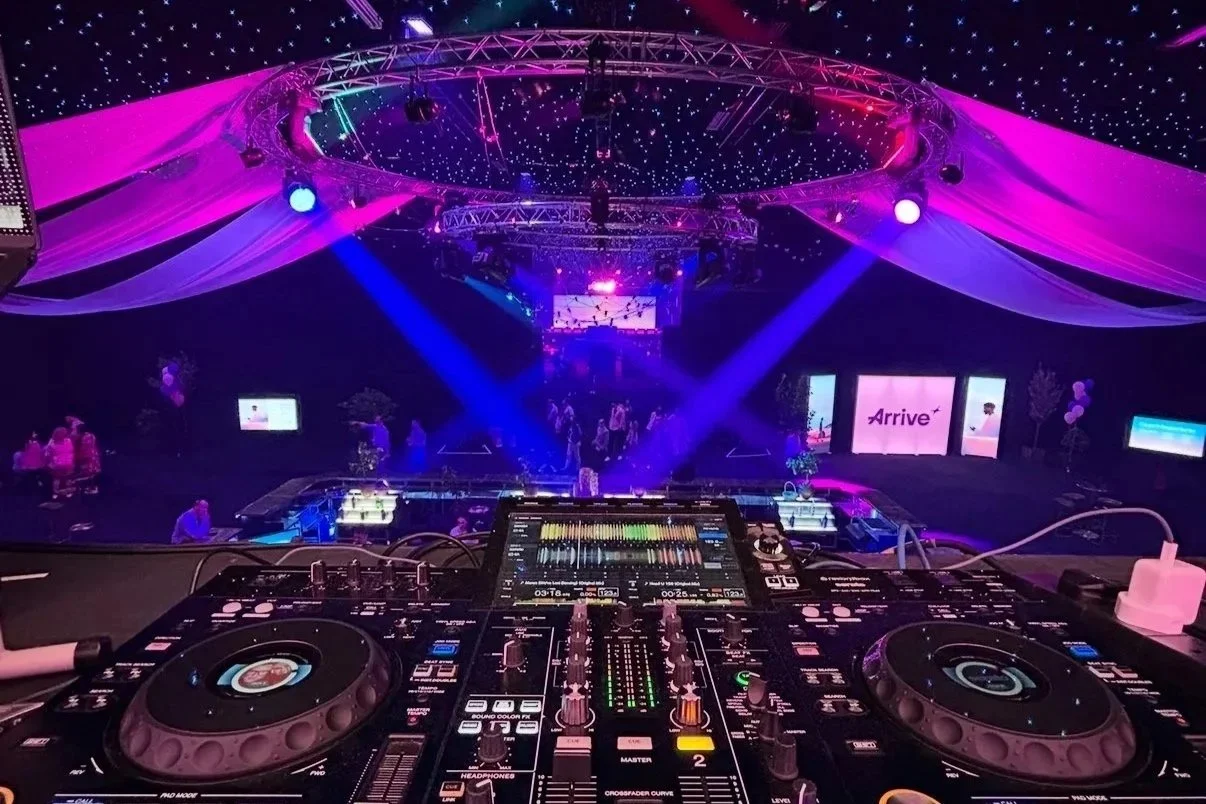 DJ equipment in the foreground inside a dance club, with colorful stage lighting, drapes, and screens displaying 'Arrive' and other visuals in the background.