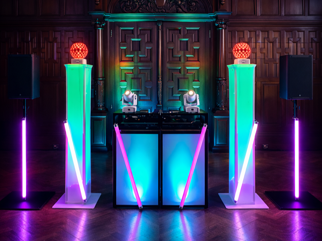Colorful DJ setup with neon lights, two large speakers, and a wooden-paneled wall in the background.