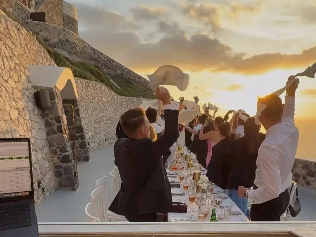 People celebrating with hats at an outdoor sunset dinner party on a terrace with stone walls.