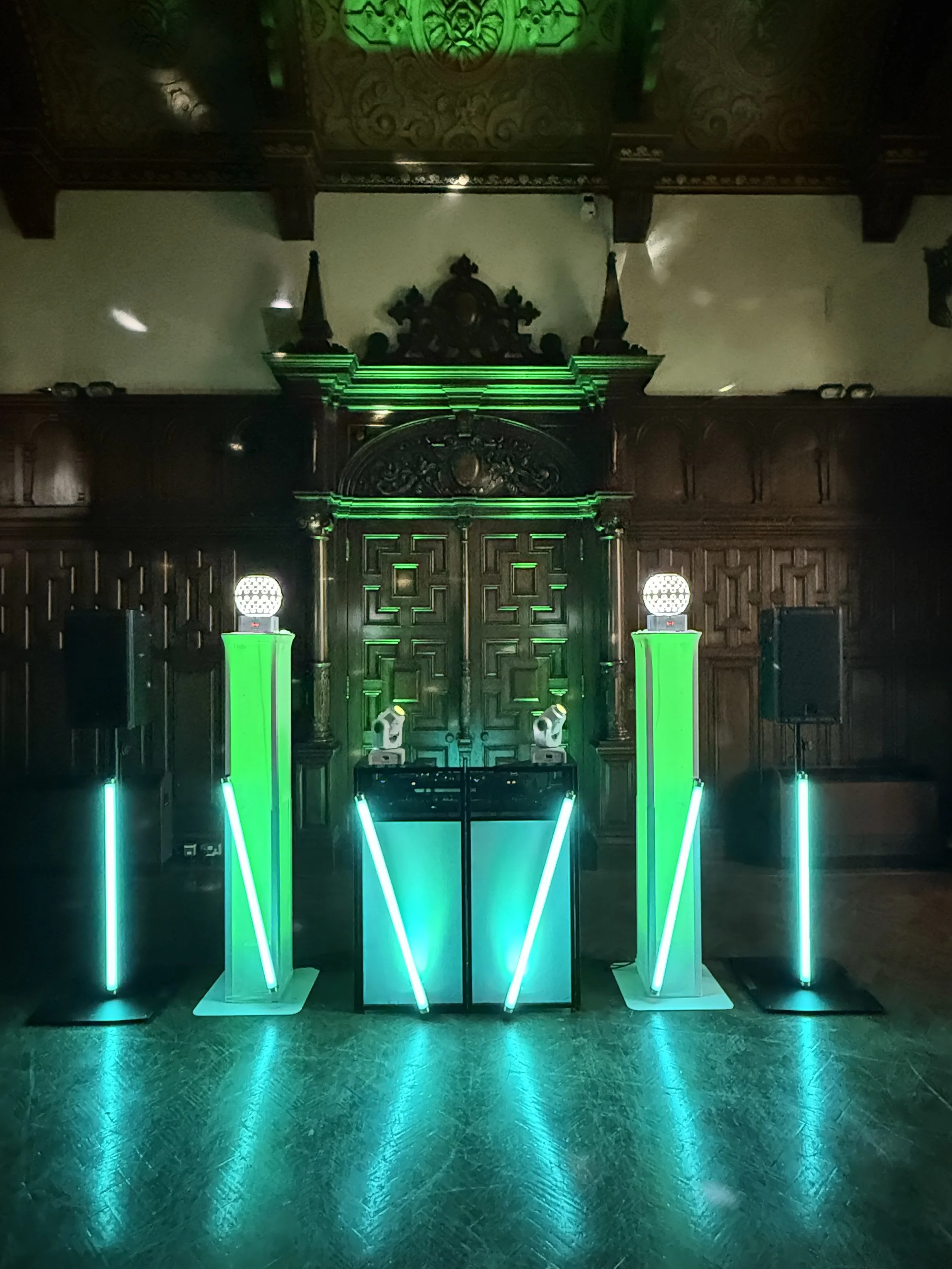 A stage with vertical and diagonal bright teal neon lights, set against intricately carved wooden wall panels with green lighting. The stage has speakers and a DJ booth.