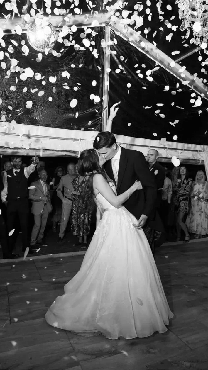 A newlywed couple dances closely at their wedding reception, surrounded by guests, with confetti falling from the ceiling and a disco ball overhead in a decorated indoor venue.