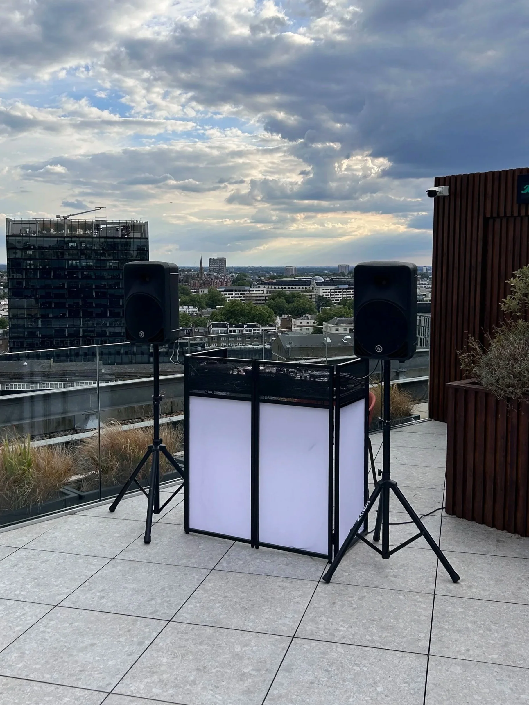 Rooftop with speakers and DJ equipment overlooking a city skyline with modern buildings, trees, and cloudy sky during daytime.